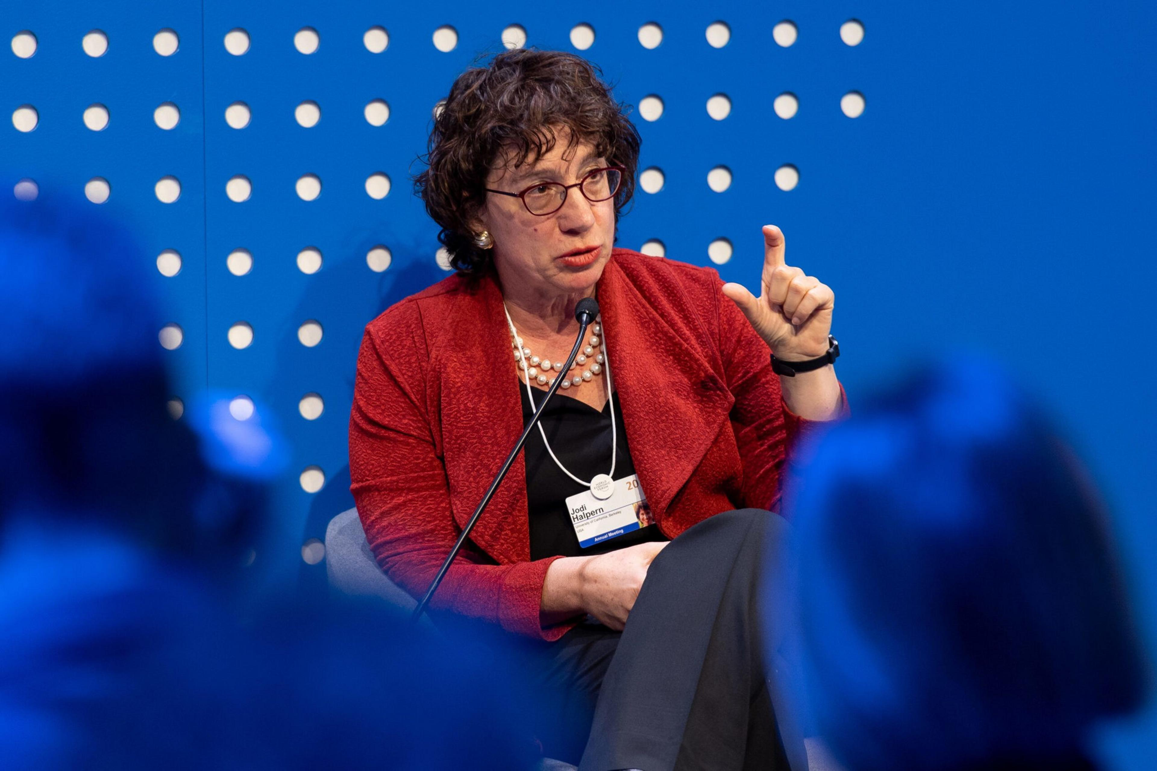 A woman with curly hair and glasses, wearing a red jacket and pearl necklace, gestures with her hand while speaking into a microphone.