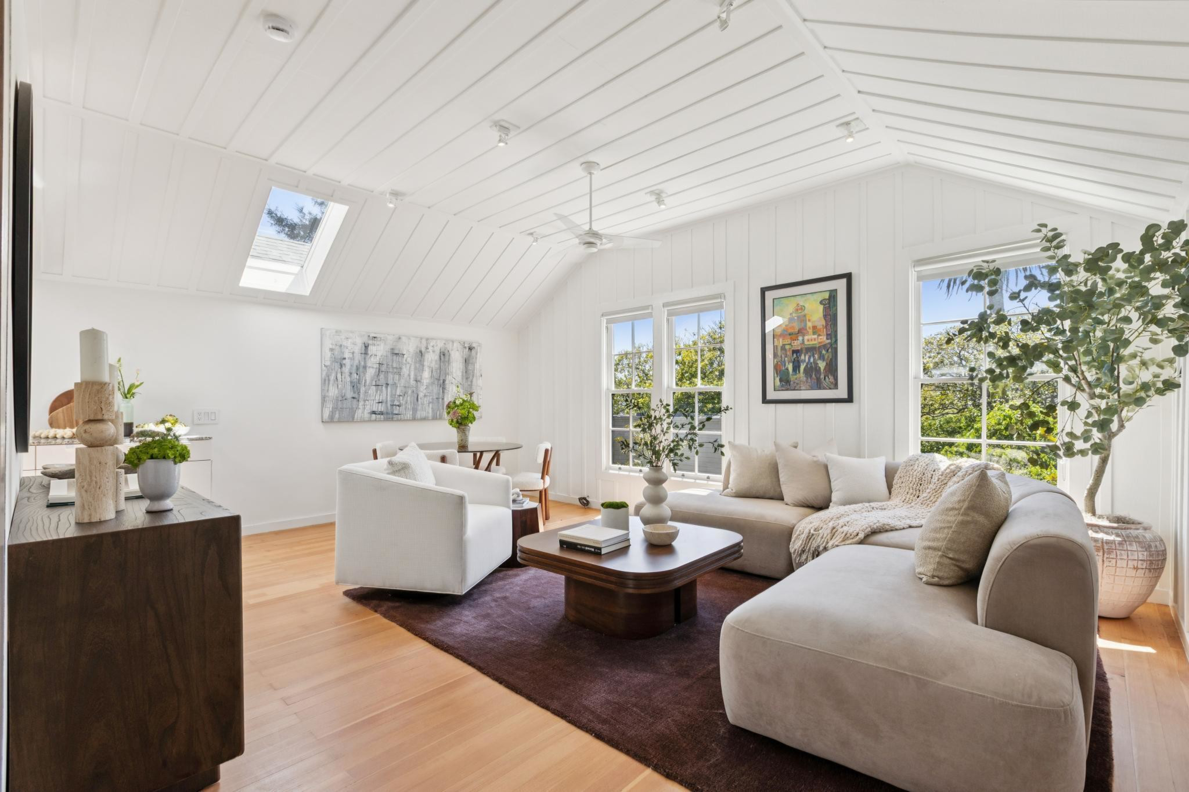 A bright living room with a beige sectional sofa, white armchair, wooden coffee table, dark rug, large potted plant, and framed artwork on white paneled walls.