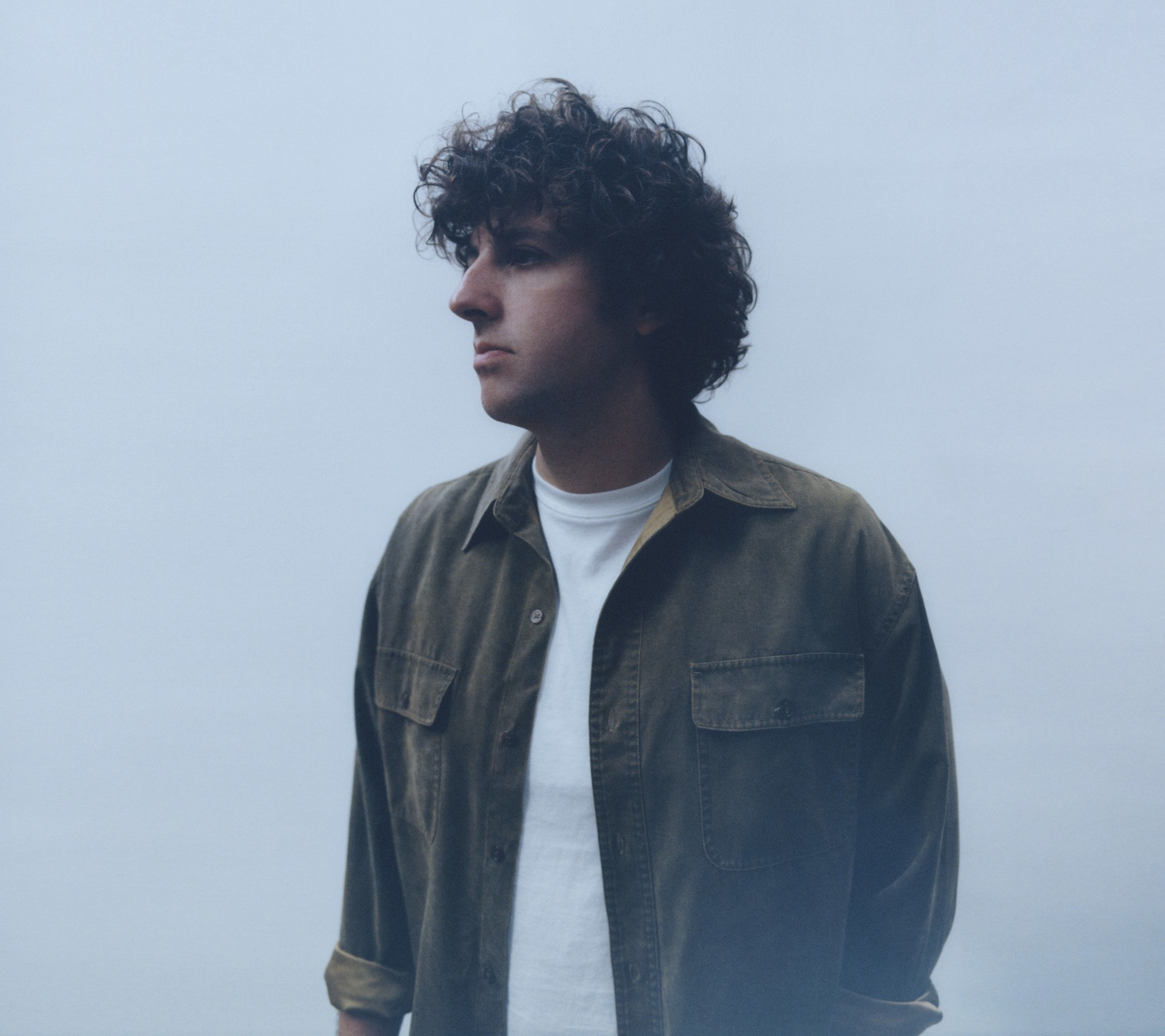 A person with curly hair looks to the left, wearing a white T-shirt under a greenish-brown jacket, set against a plain, light background.