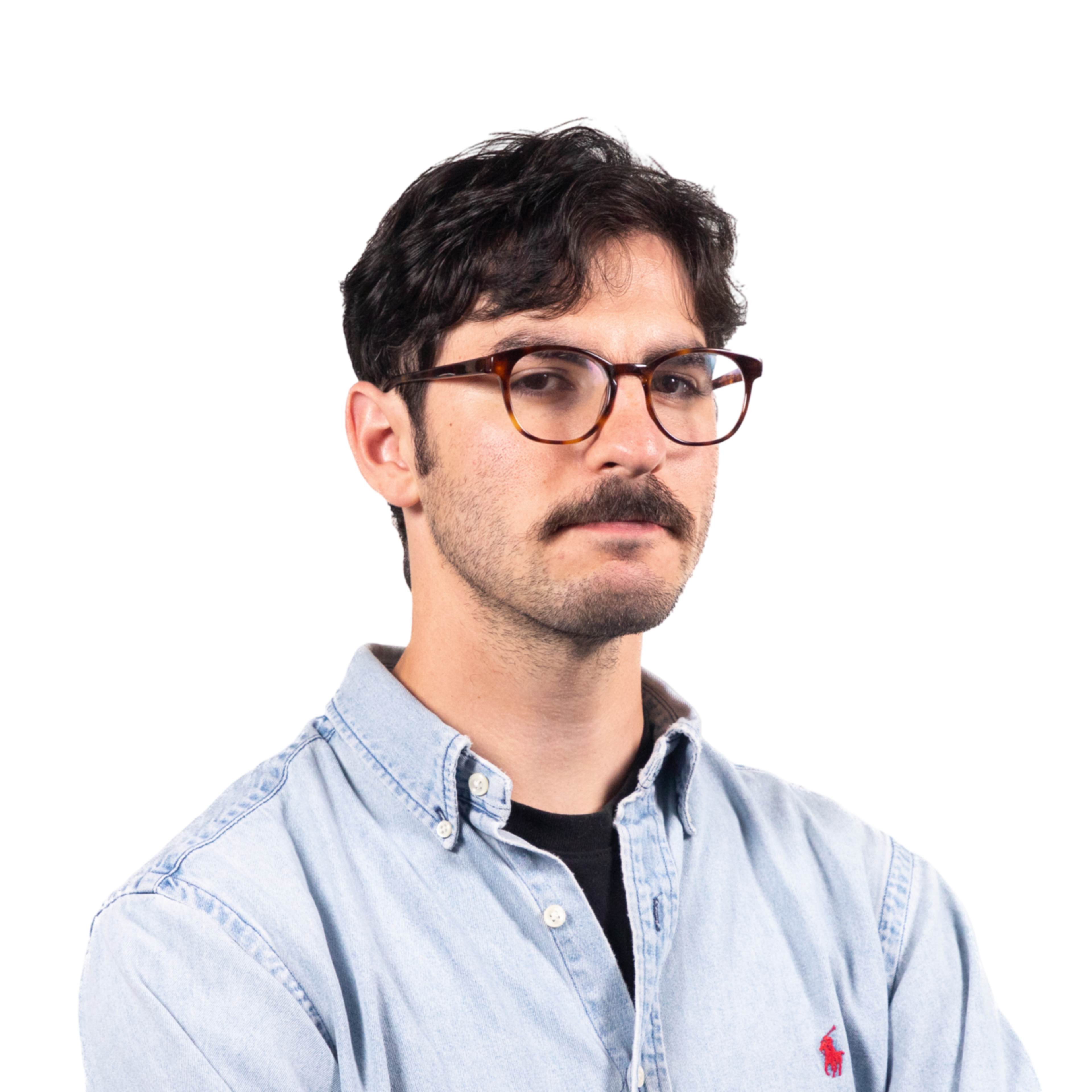 A man with dark wavy hair, glasses, and a mustache wears a light blue denim shirt over a black T-shirt, looking forward with a neutral expression.