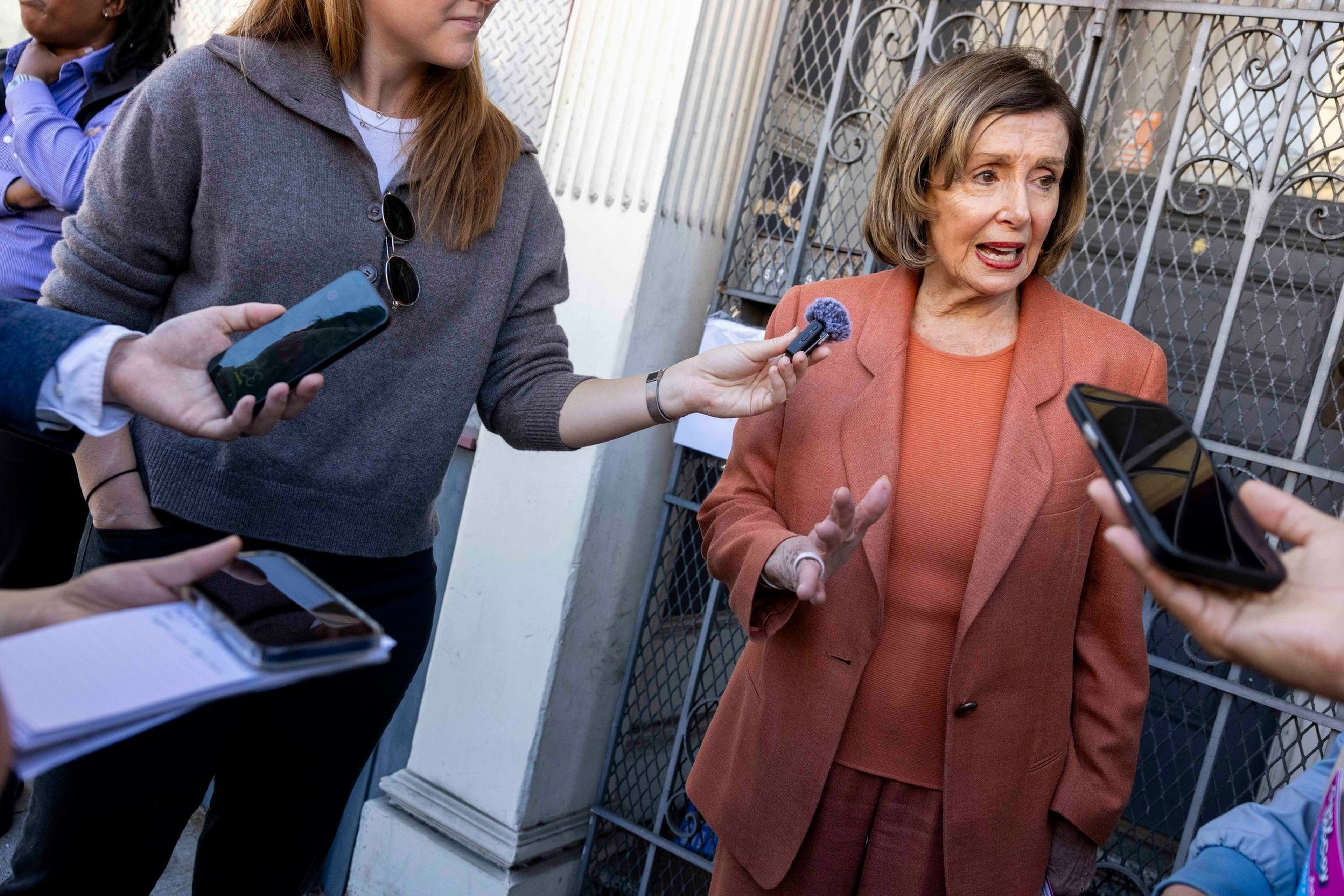 A woman in a rust-colored suit is talking while several people hold out microphones and phones to record her outside by a metal gate.