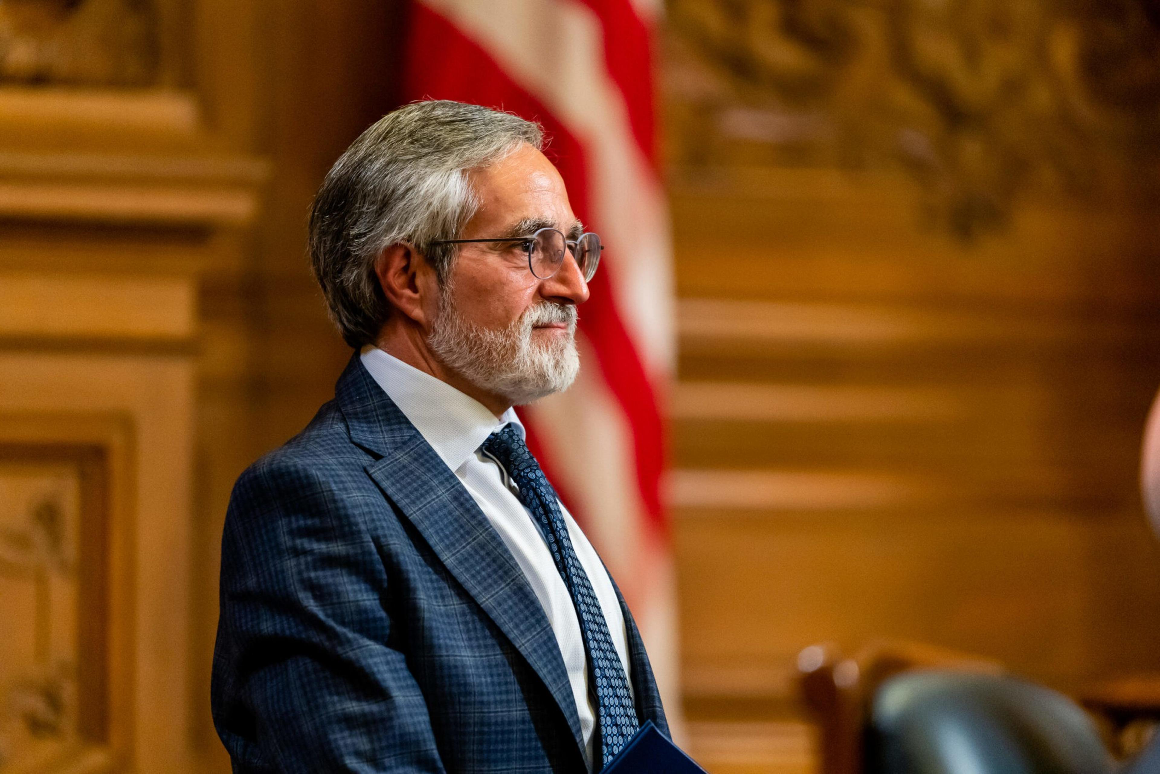 An older man with gray hair, a beard, and glasses stands in a suit and tie, with a blurred American flag and ornate wooden wall behind him.