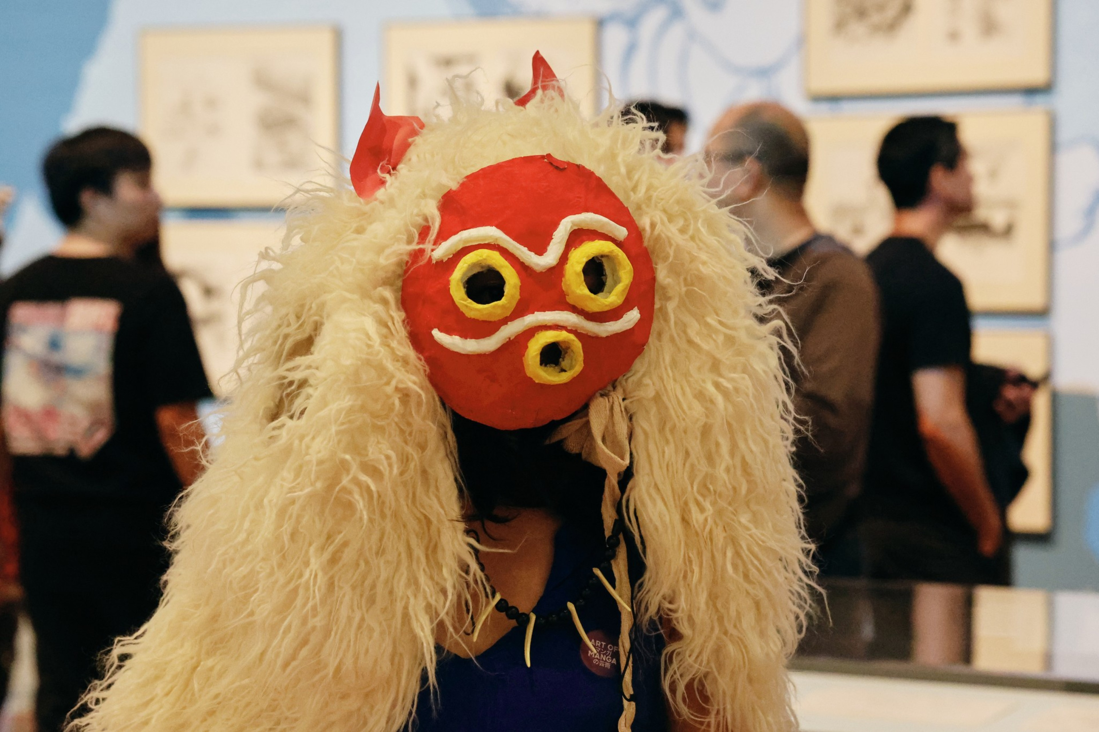 A person wears a red mask with yellow-rimmed eyes and mouth and a fluffy beige wig with two small red horns, standing in an art gallery.