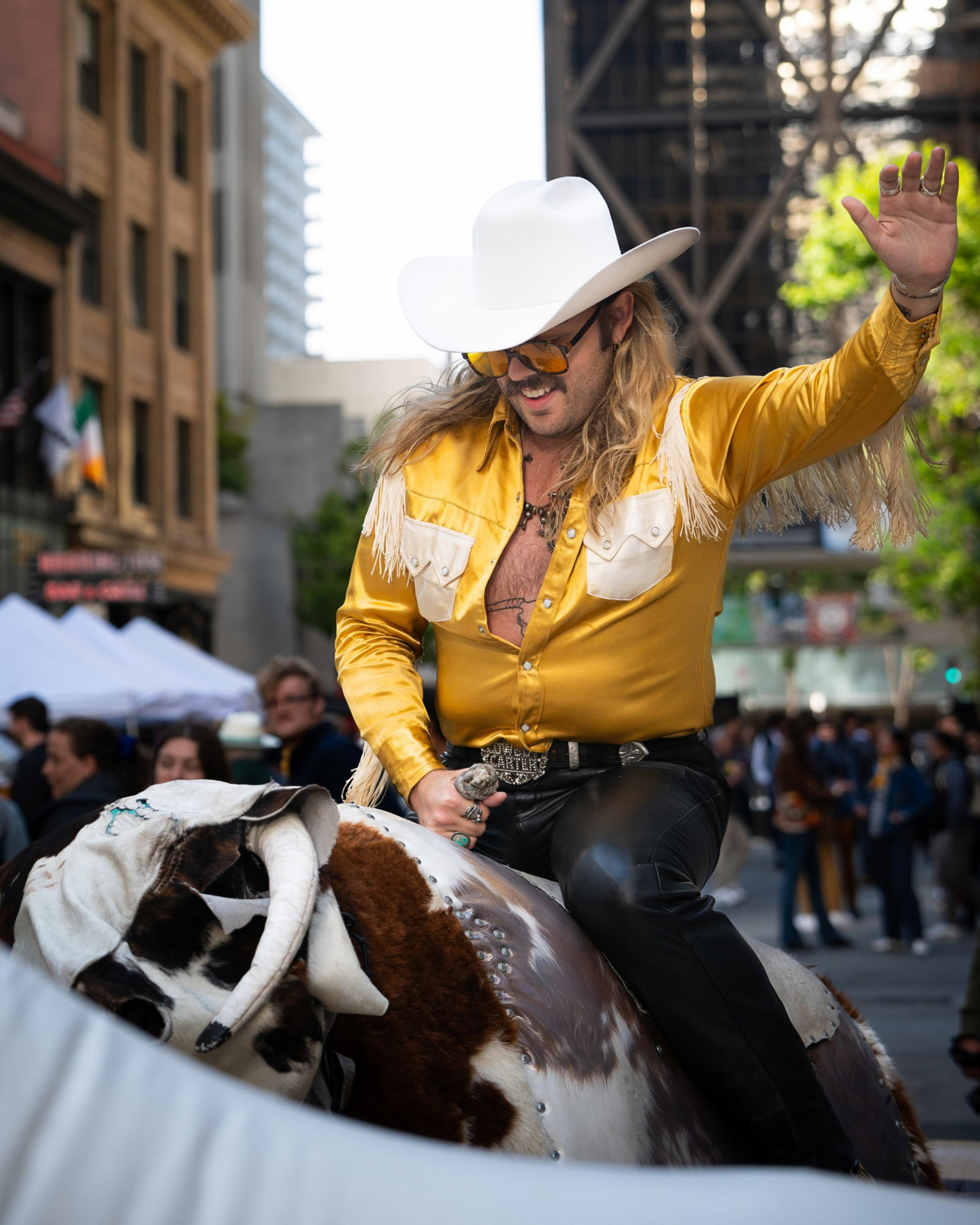 A man in a yellow fringed shirt, white cowboy hat, and sunglasses rides a mechanical bull in a city street with people watching behind him.