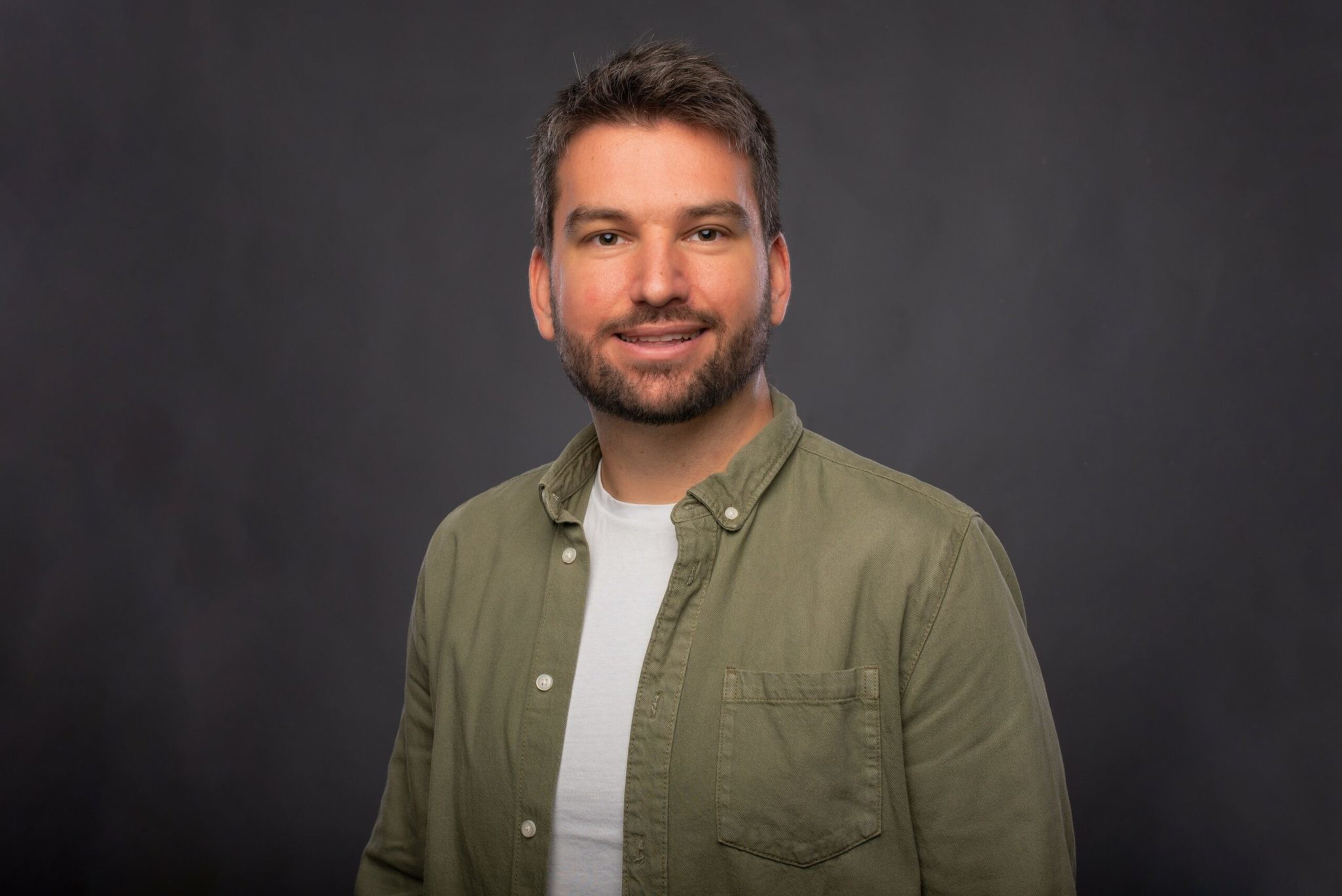A man with short dark hair and a beard smiles, wearing a white t-shirt under an olive green button-up shirt, standing against a dark background.