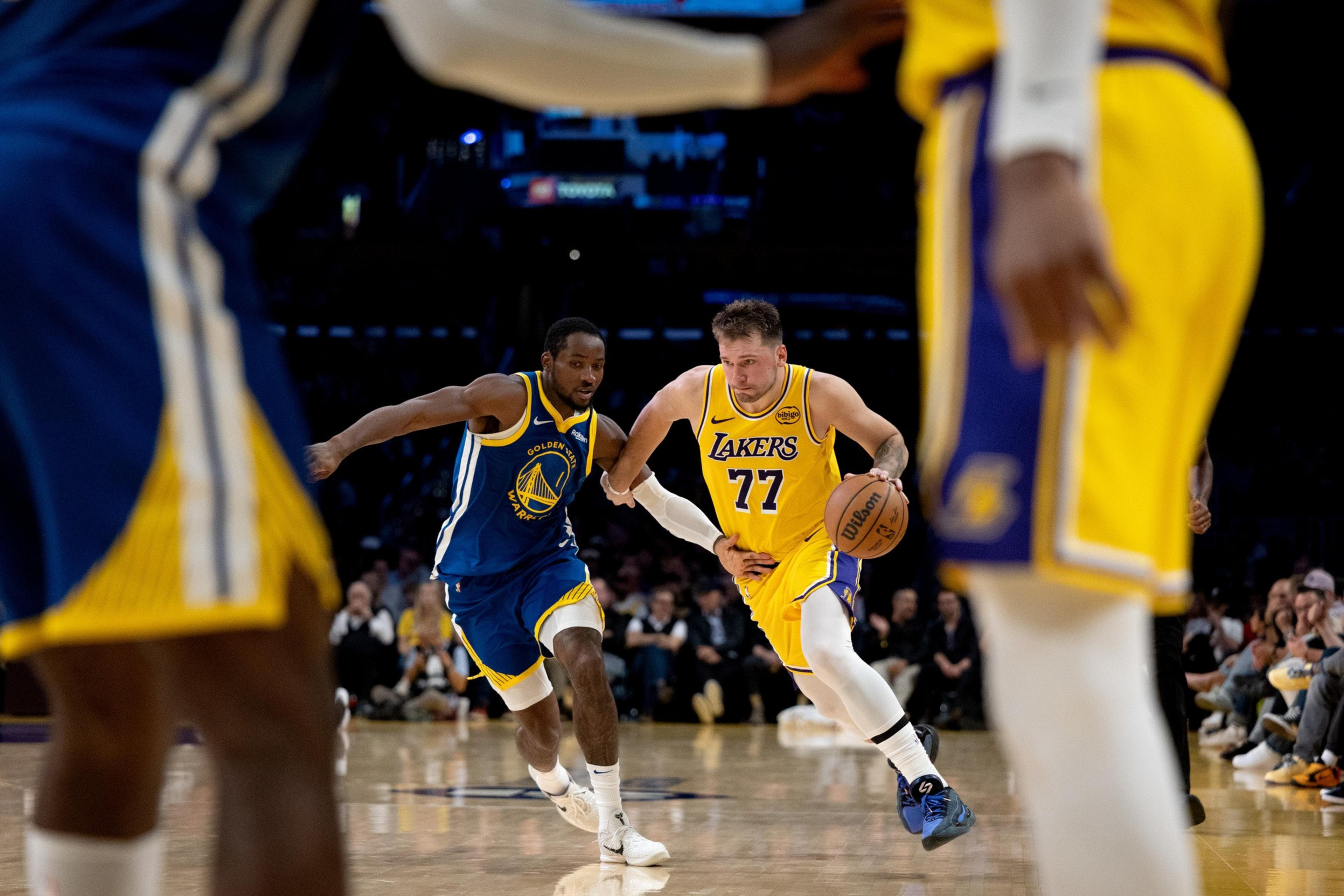 A Lakers player, wearing jersey number 77, dribbles the basketball while being closely guarded by a Golden State Warriors player during a game.