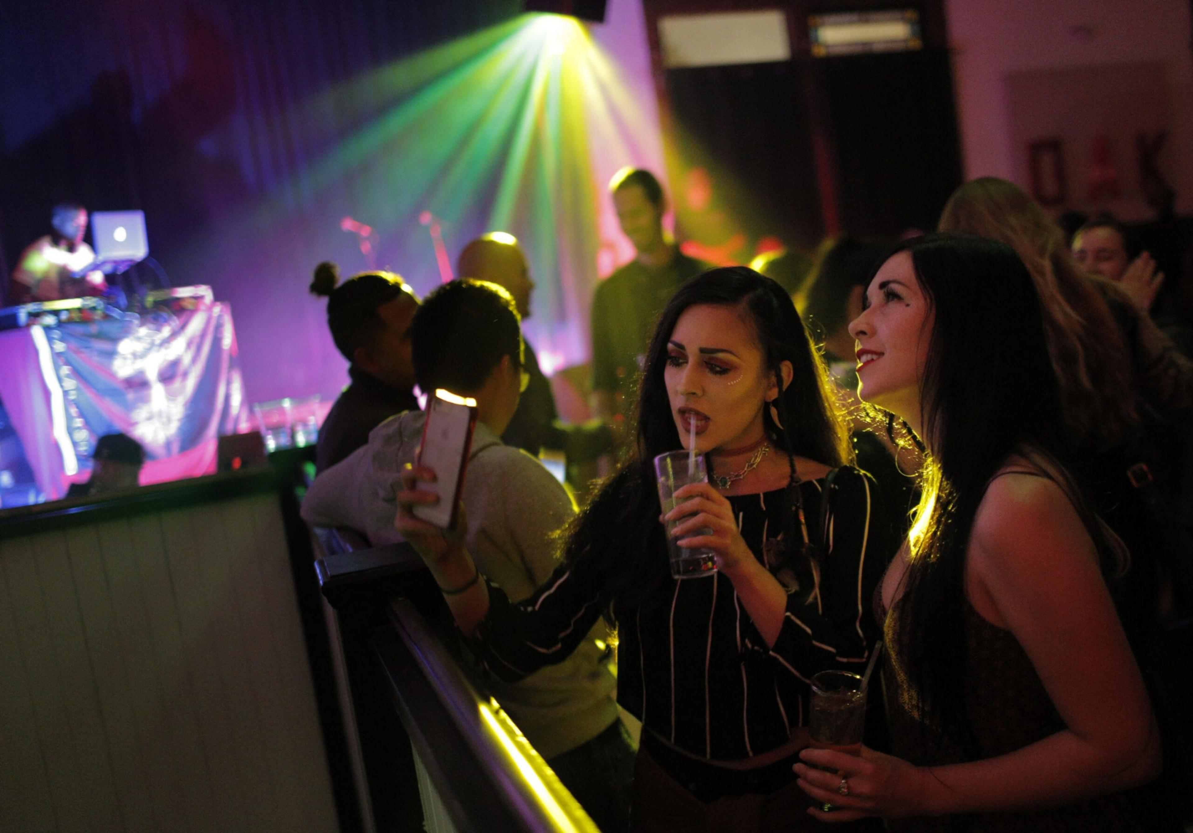 Two women hold drinks and take a selfie in a dimly lit club with colorful stage lights and a DJ in the background.