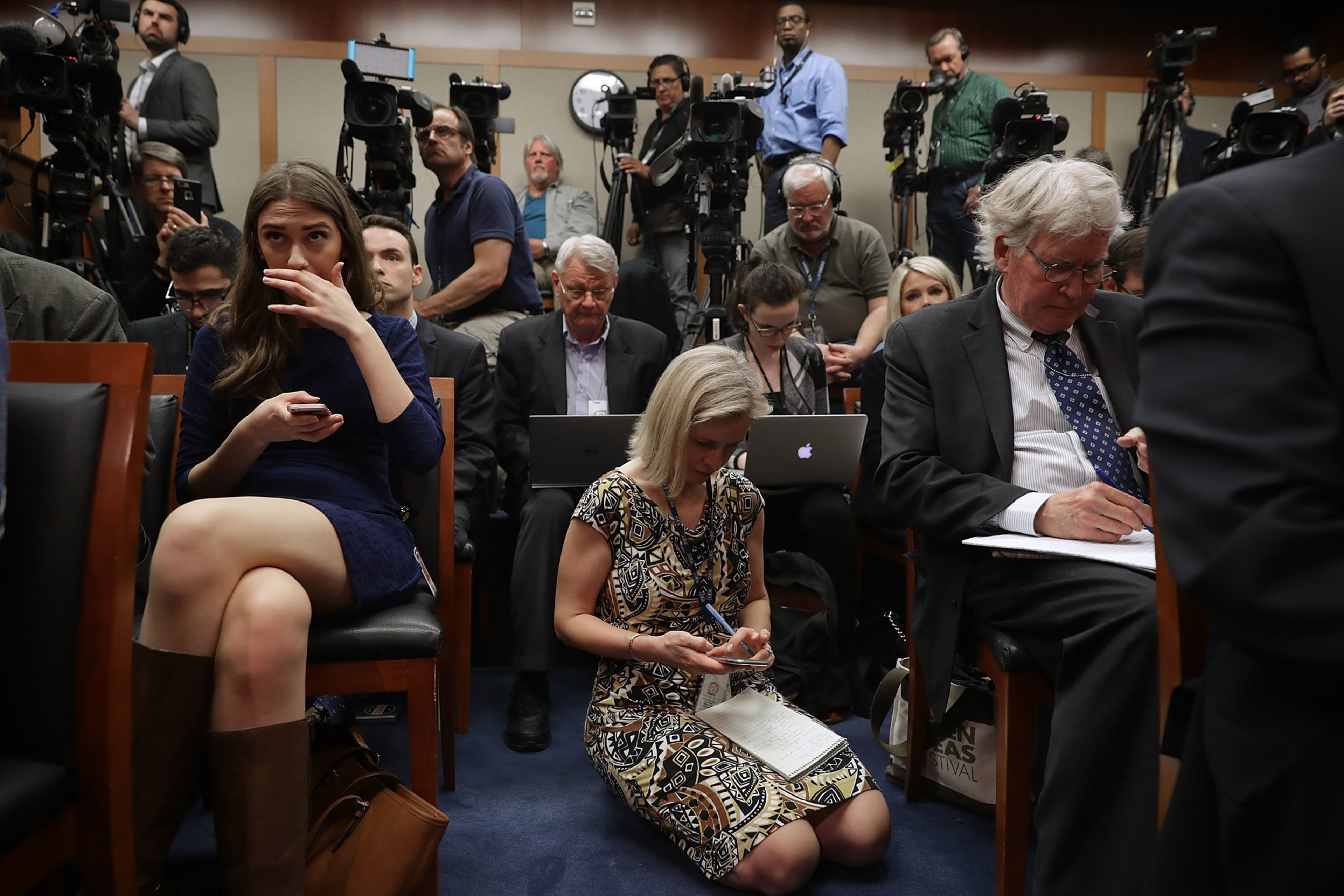 Reporters and cameramen focus intently, some seated and others standing, with laptops and cameras in a crowded room during a news event.