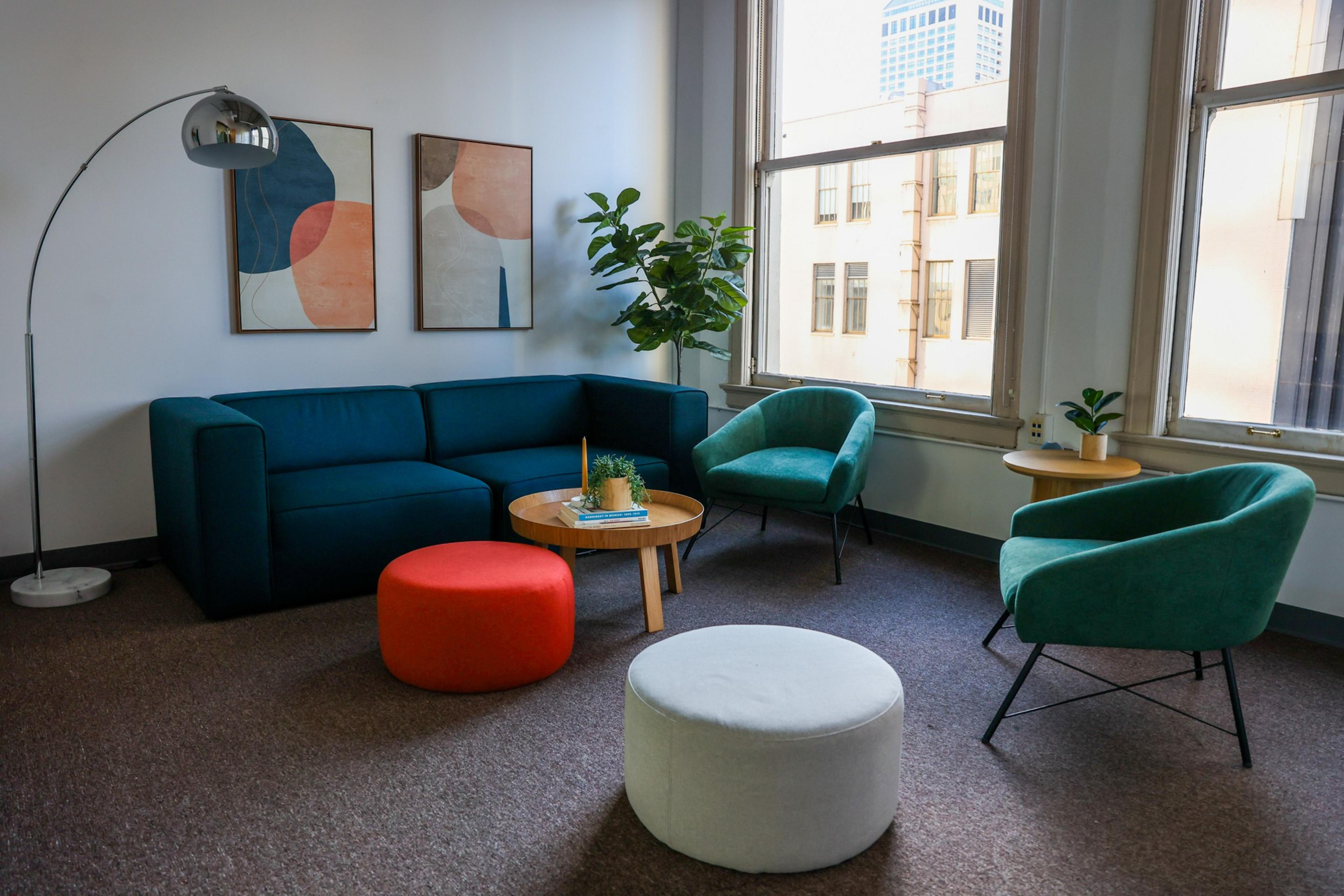 A modern living room with a dark teal sofa, two teal armchairs, a white and a red round ottoman, a round wooden coffee table, and two abstract paintings on the wall.