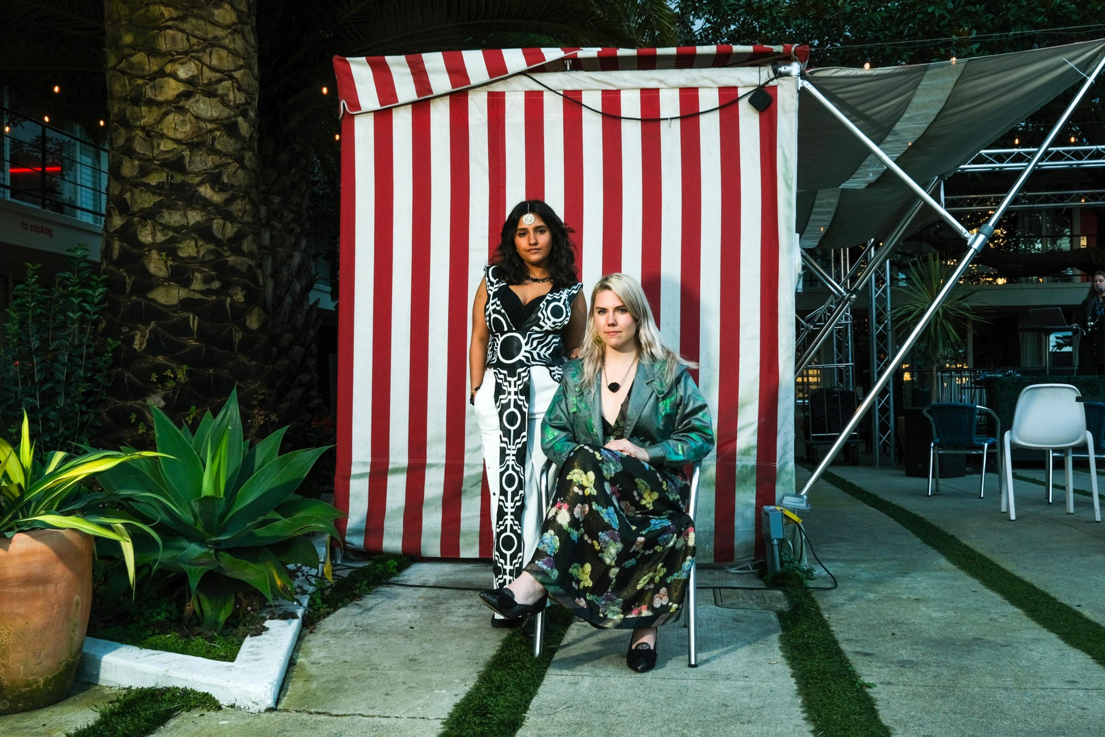 Two women pose in front of a red and white striped backdrop, one standing, the other seated, amid greenery and venue chairs.