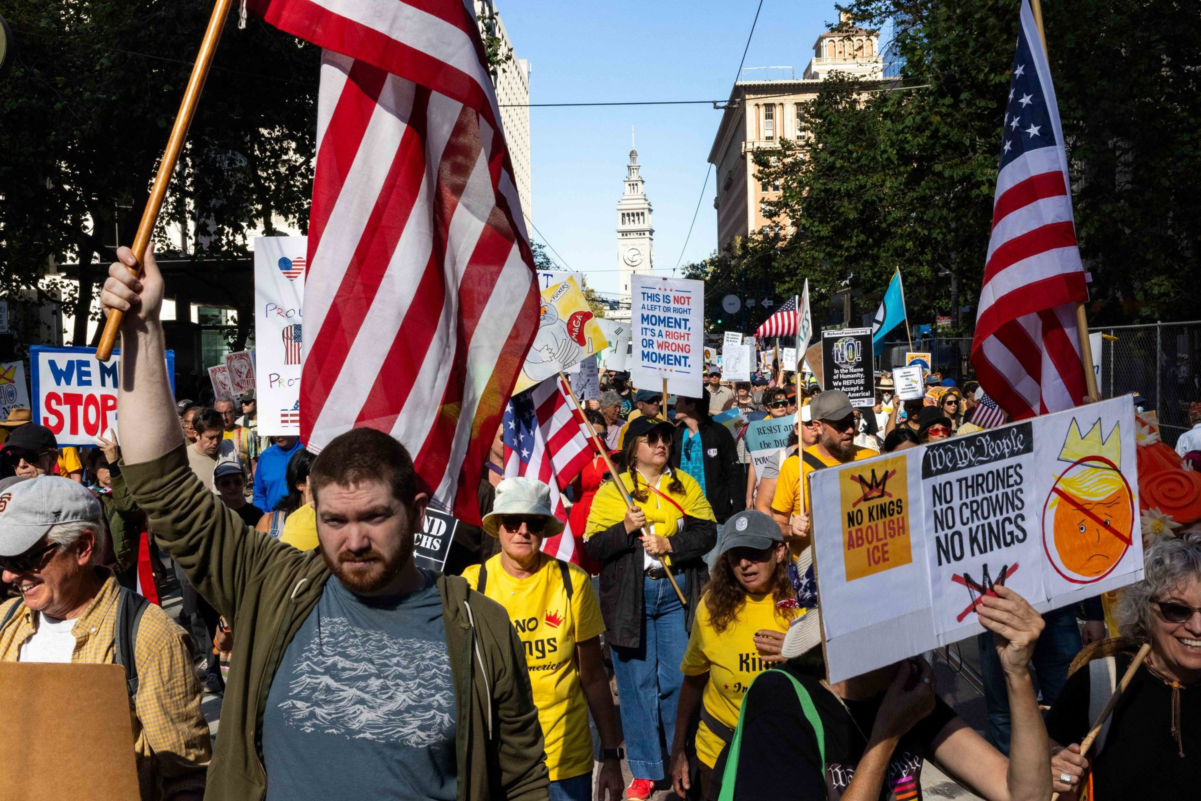 A crowd marches holding American flags and signs including “No Thrones No Crowns No Kings” and “This is not a right or wrong moment.”