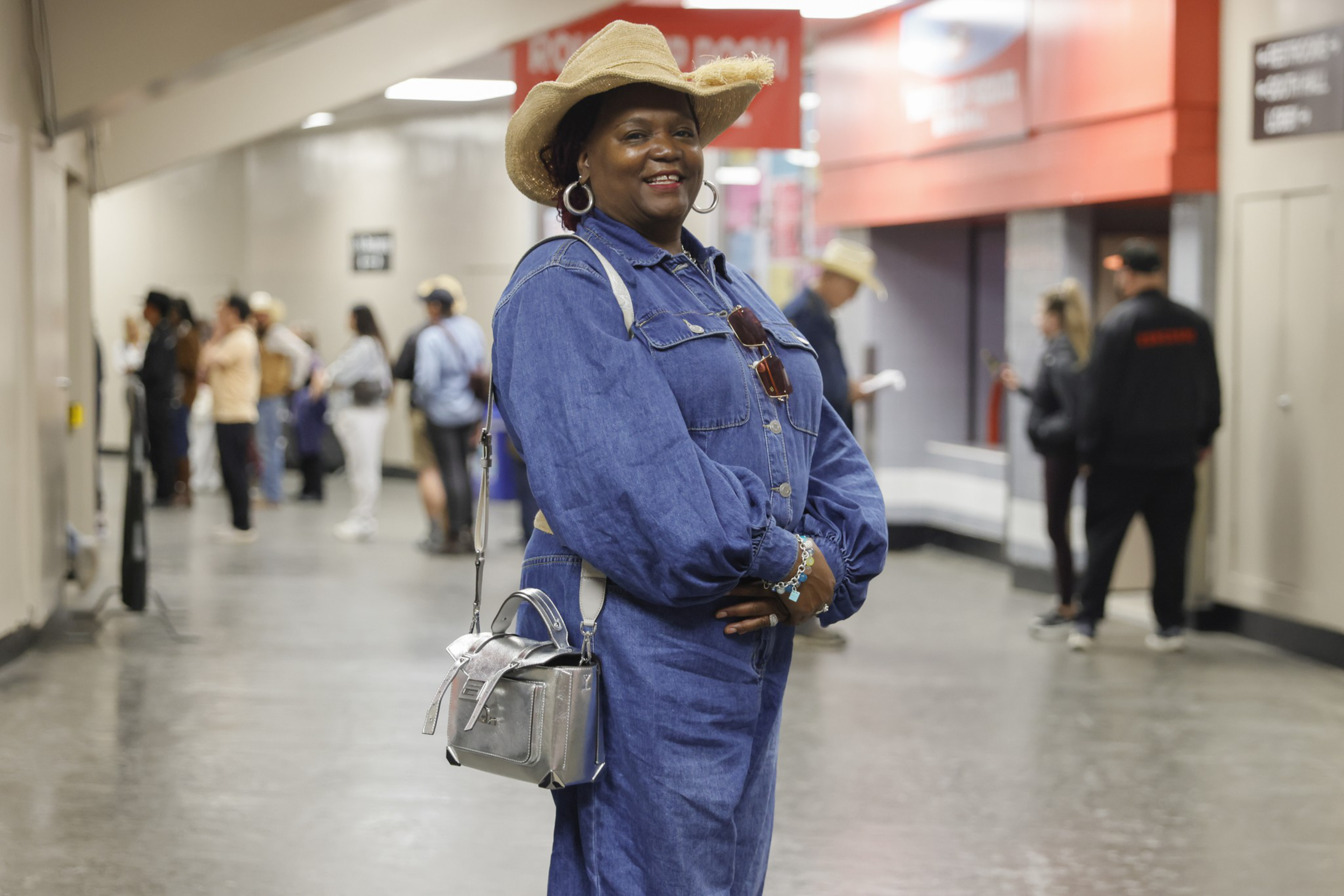 A woman in a blue denim dress and straw hat smiles while standing in a busy indoor hallway with people in the background.