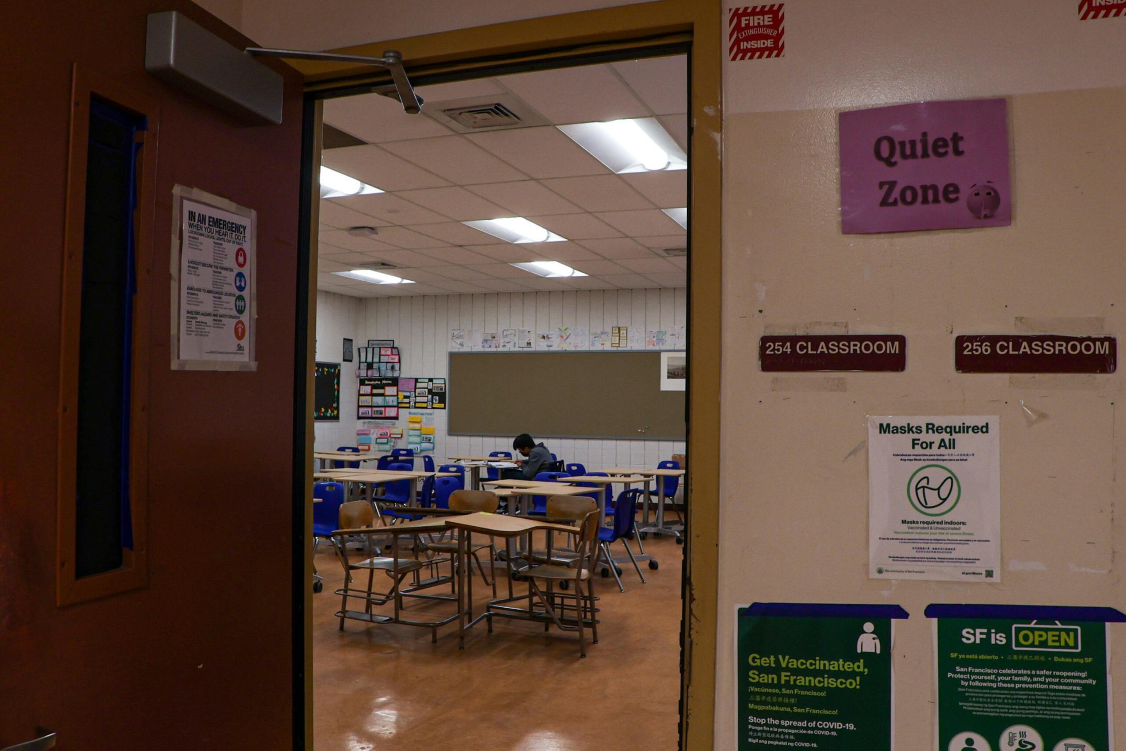 A mostly empty classroom with scattered desks, one student wearing a mask seated and working, and various signs outside the door including "Quiet Zone" and mask requirements.
