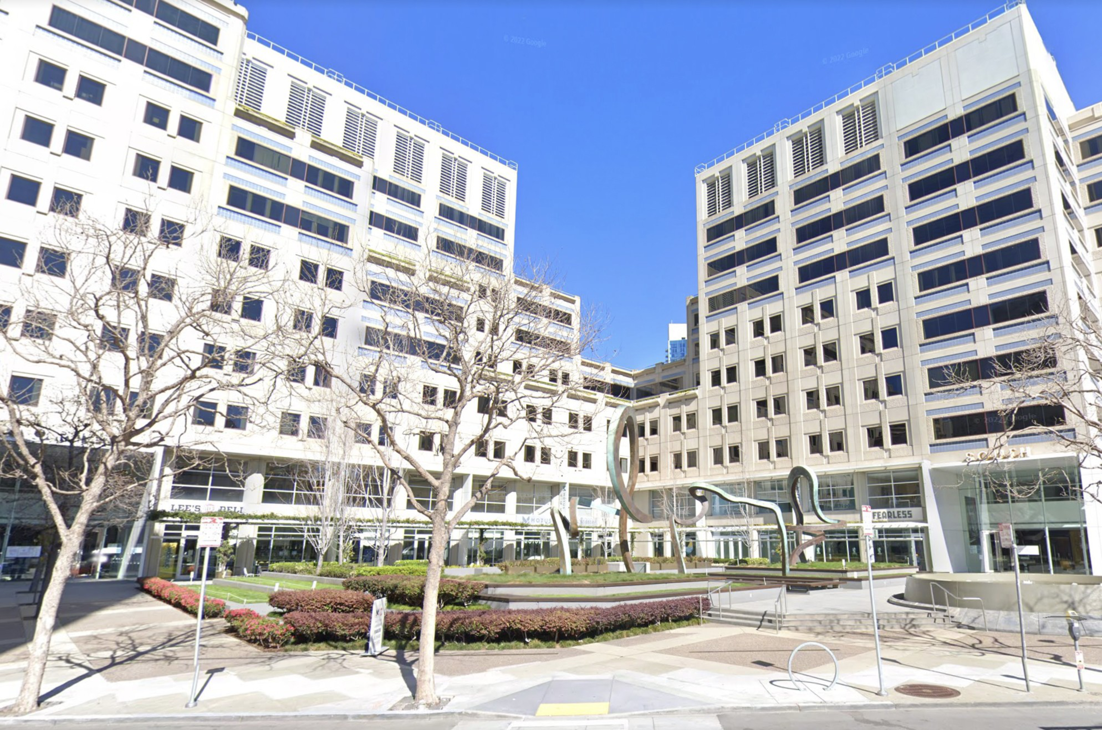 A plaza with leafless trees features modern metal sculptures and is surrounded by white multi-story office buildings under a clear blue sky.