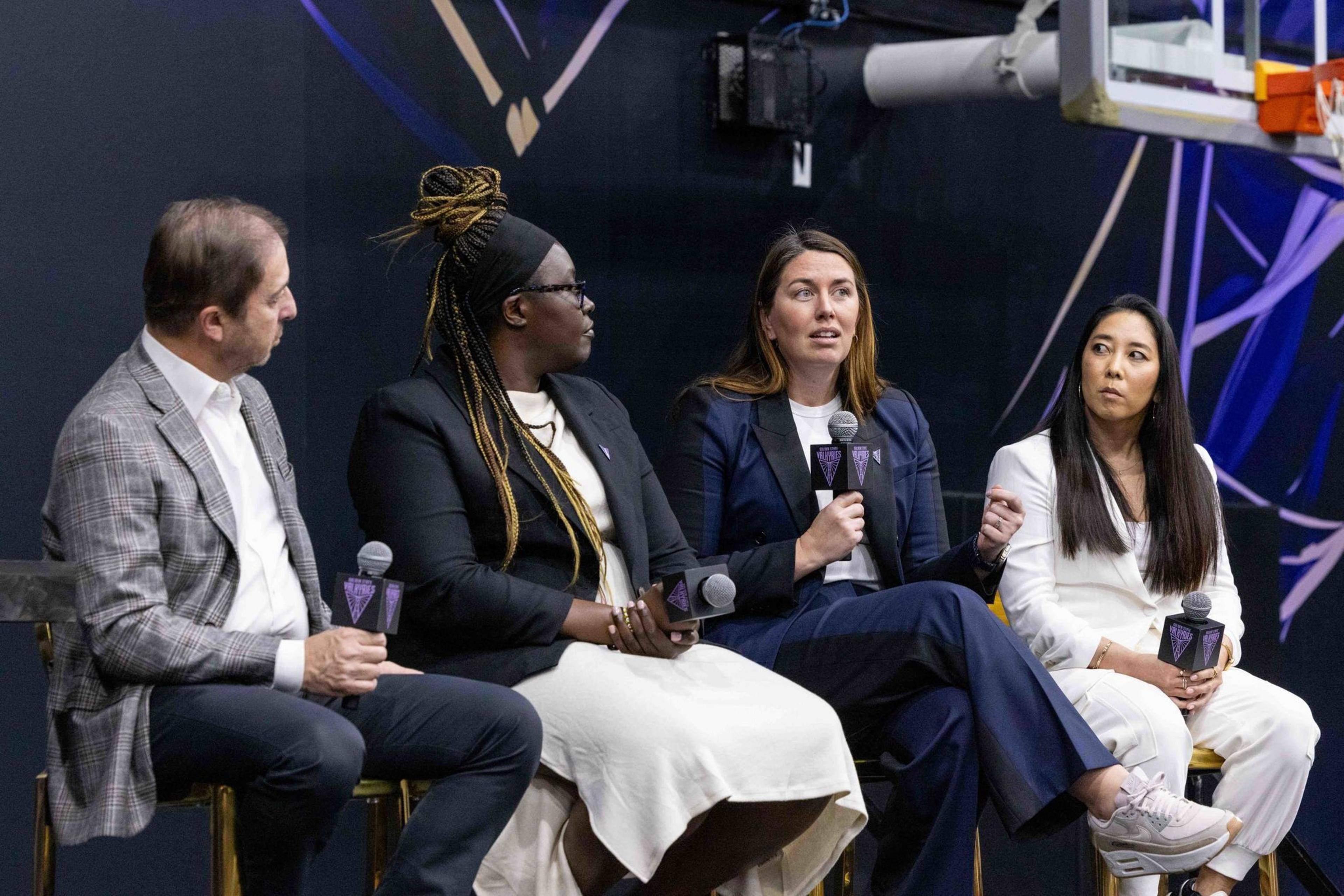 Four individuals are seated on a panel, each holding a microphone. They're engaged in a discussion in front of a background featuring a basketball hoop.