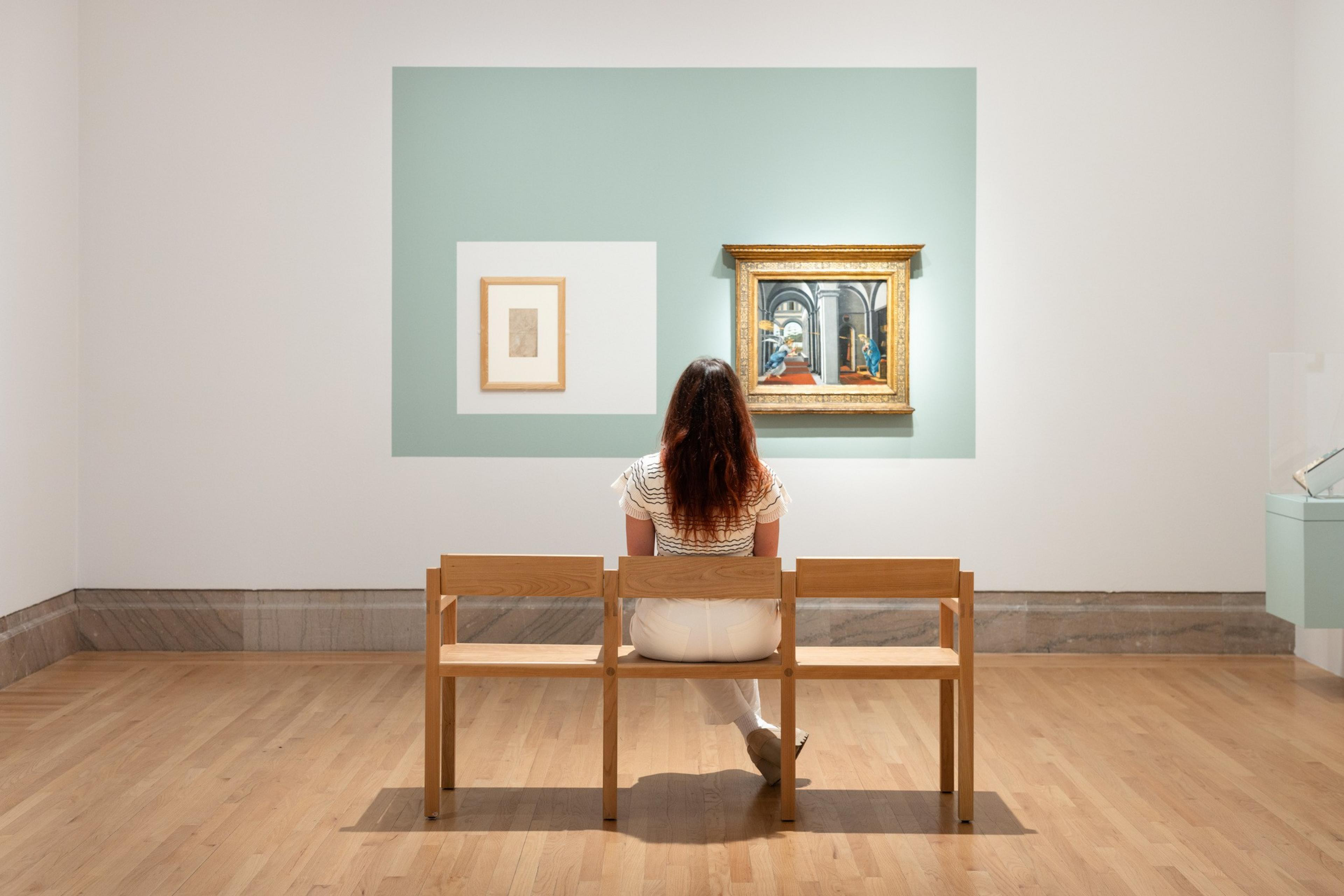 A woman in white pants and a striped short sleeve top sits on a wooden bench in an art gallery looking at a painting.