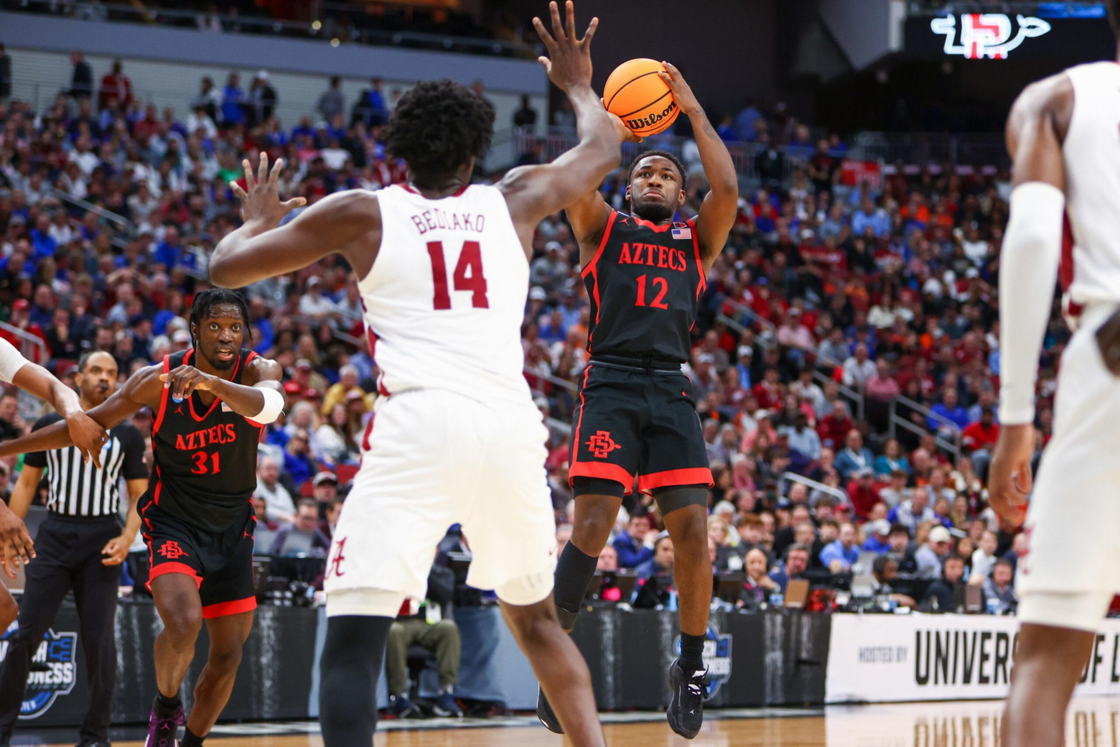 A basketball player wearing jersey number 12 from the Aztecs team jumps to shoot the ball while a defender wearing number 14 raises his arms in defense.