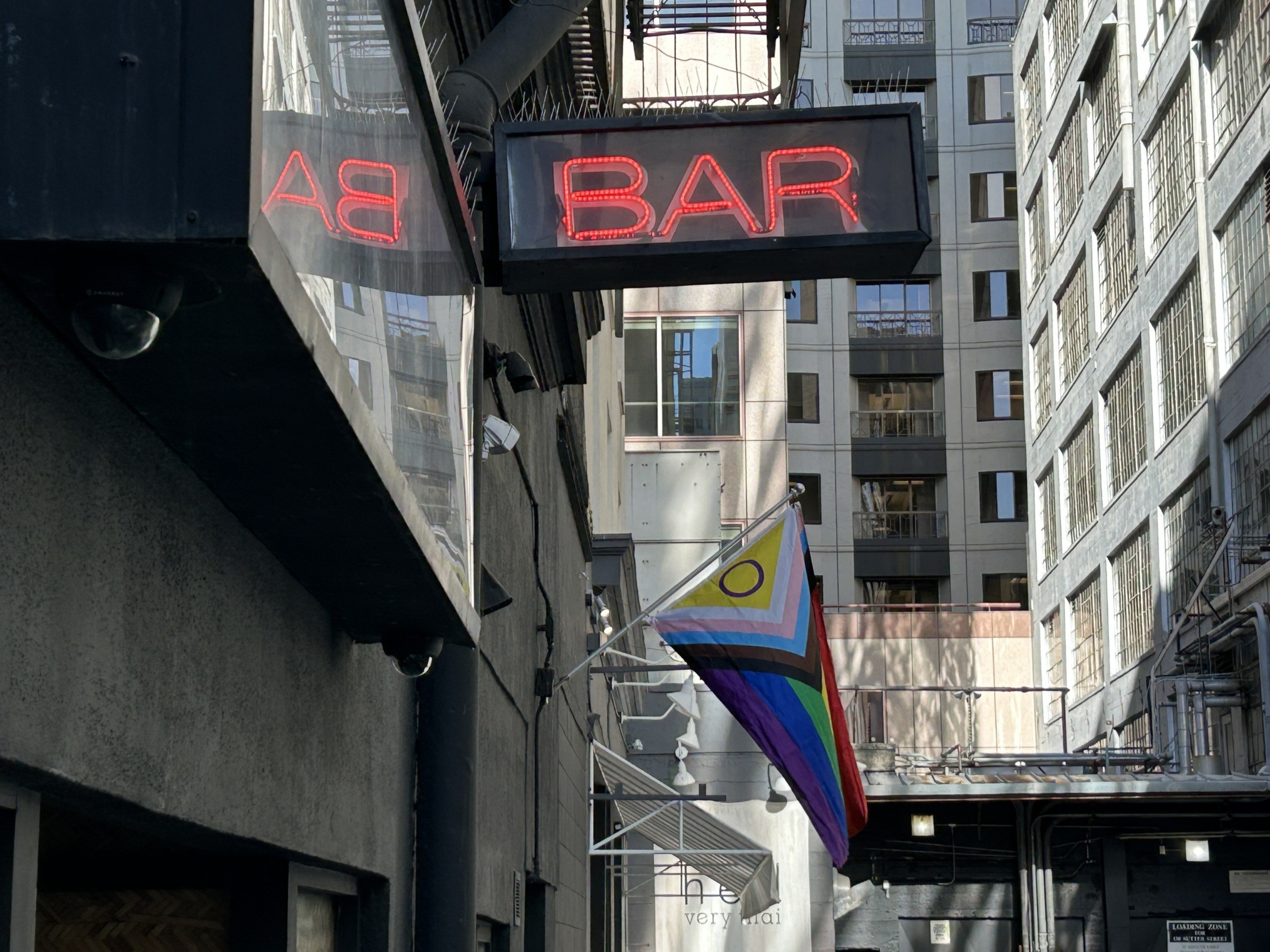 A neon red "BAR" sign hangs above a narrow alley with a Progress Pride flag flying, surrounded by tall buildings with many windows.