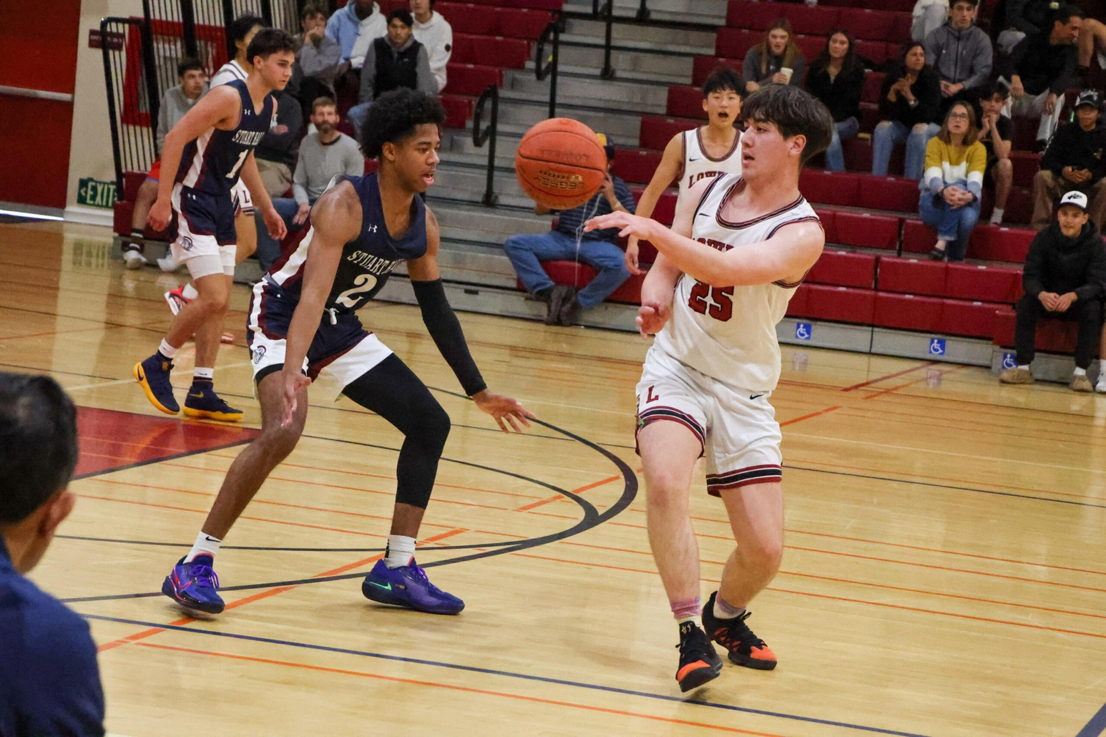 A basketball player in white passes the ball while a defensive player in dark blue reaches to block him on a gym court with spectators seated behind.