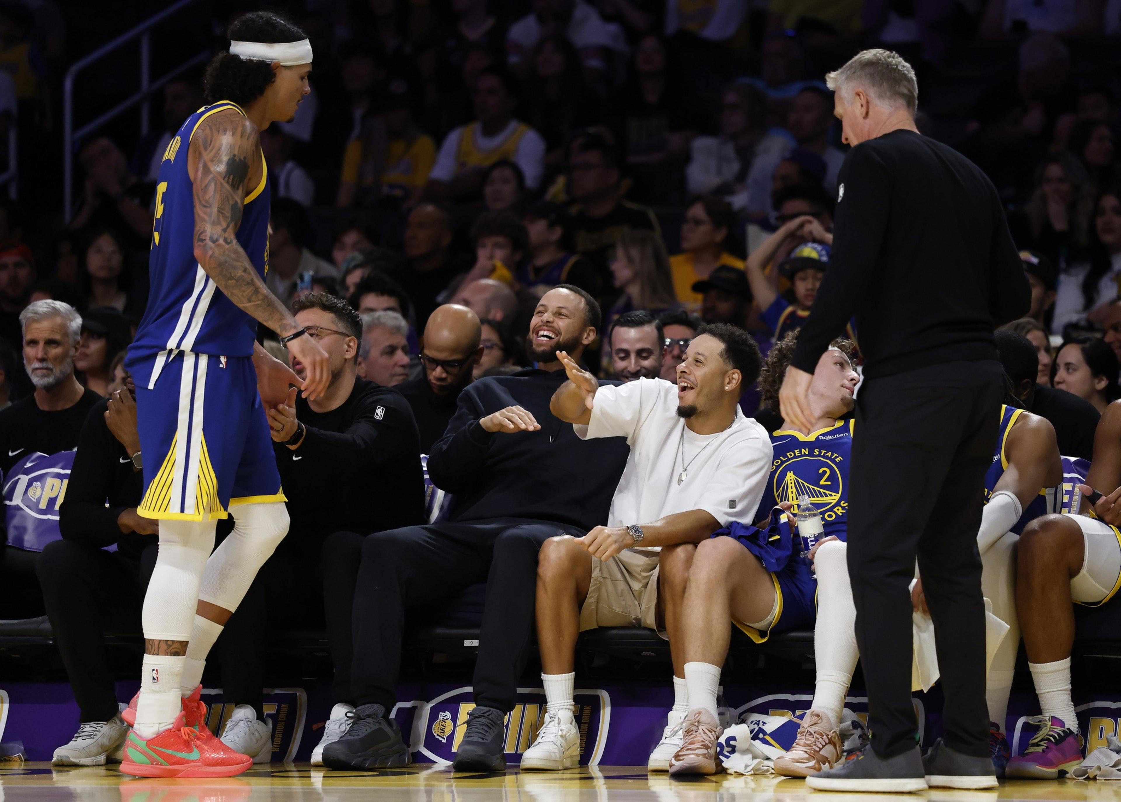 A basketball player in a blue and yellow uniform approaches a laughing group of seated men, while a standing man in black watches nearby.