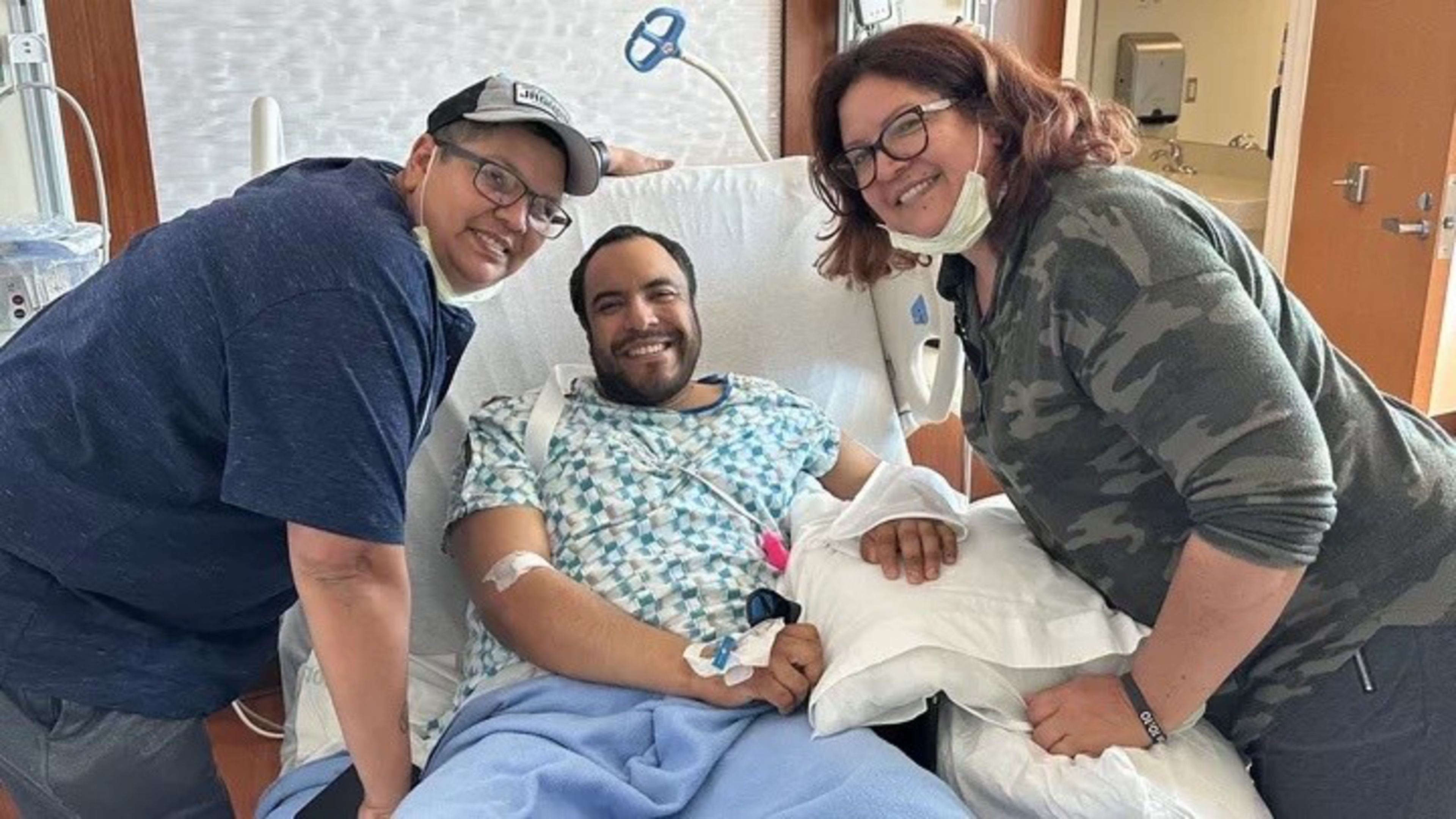 A smiling man lies in a hospital bed wearing a gown, with two people leaning in close beside him, all appearing happy and supportive.