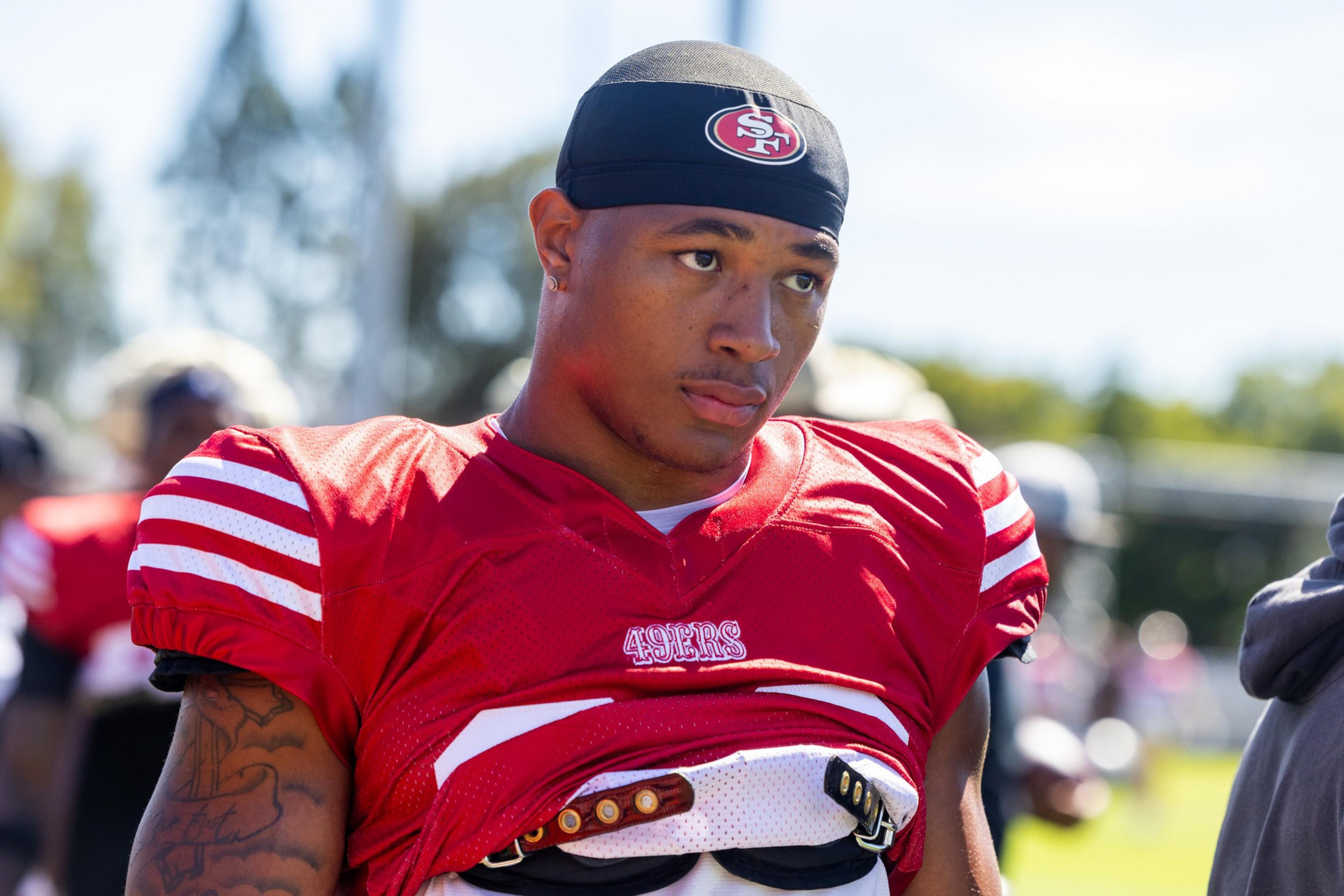 A football player wearing a red San Francisco 49ers jersey and headband looks to the side during practice under a sunny sky.