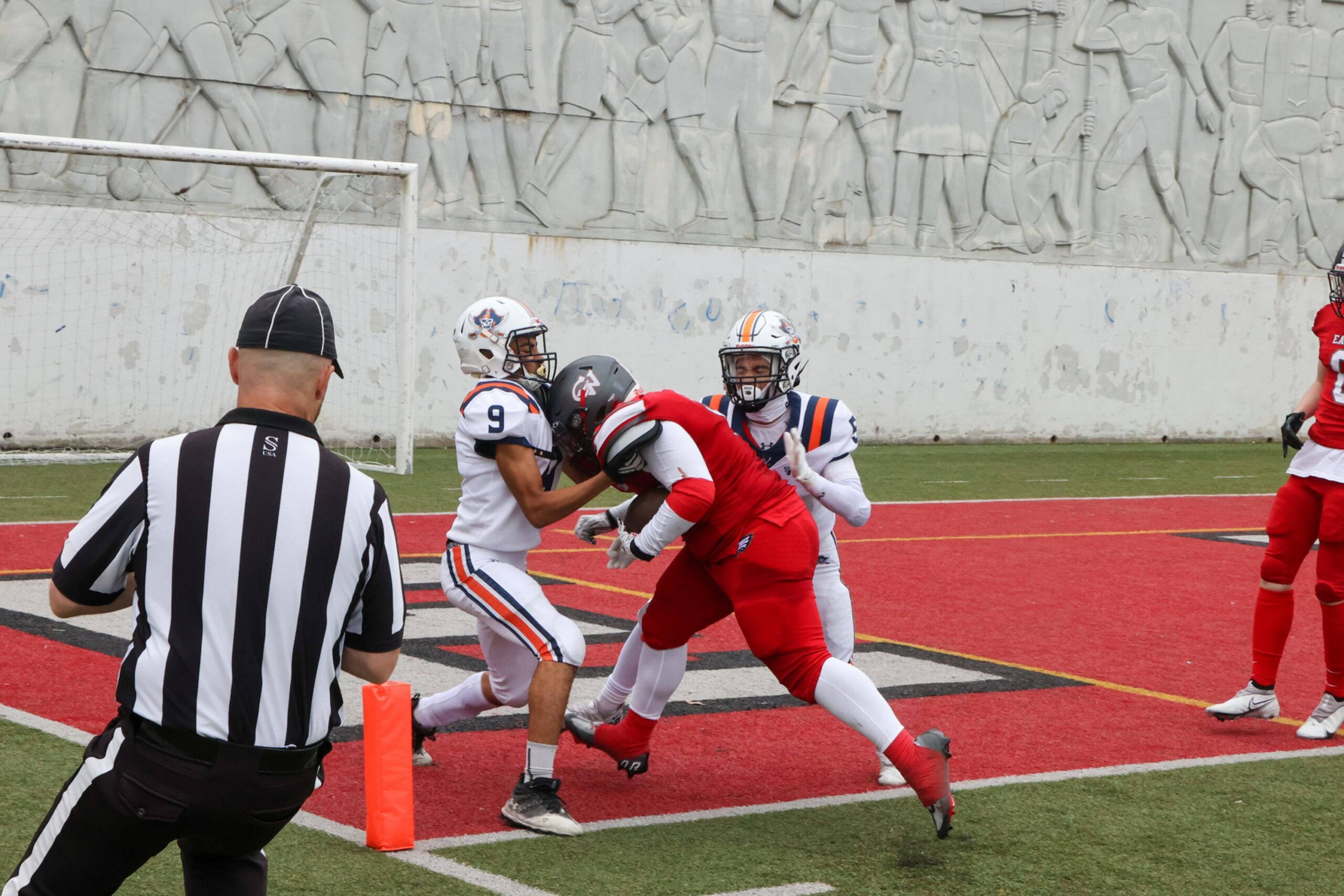 A football player in a red uniform tackles a player in a white uniform near the goal line, while a referee watches closely nearby.