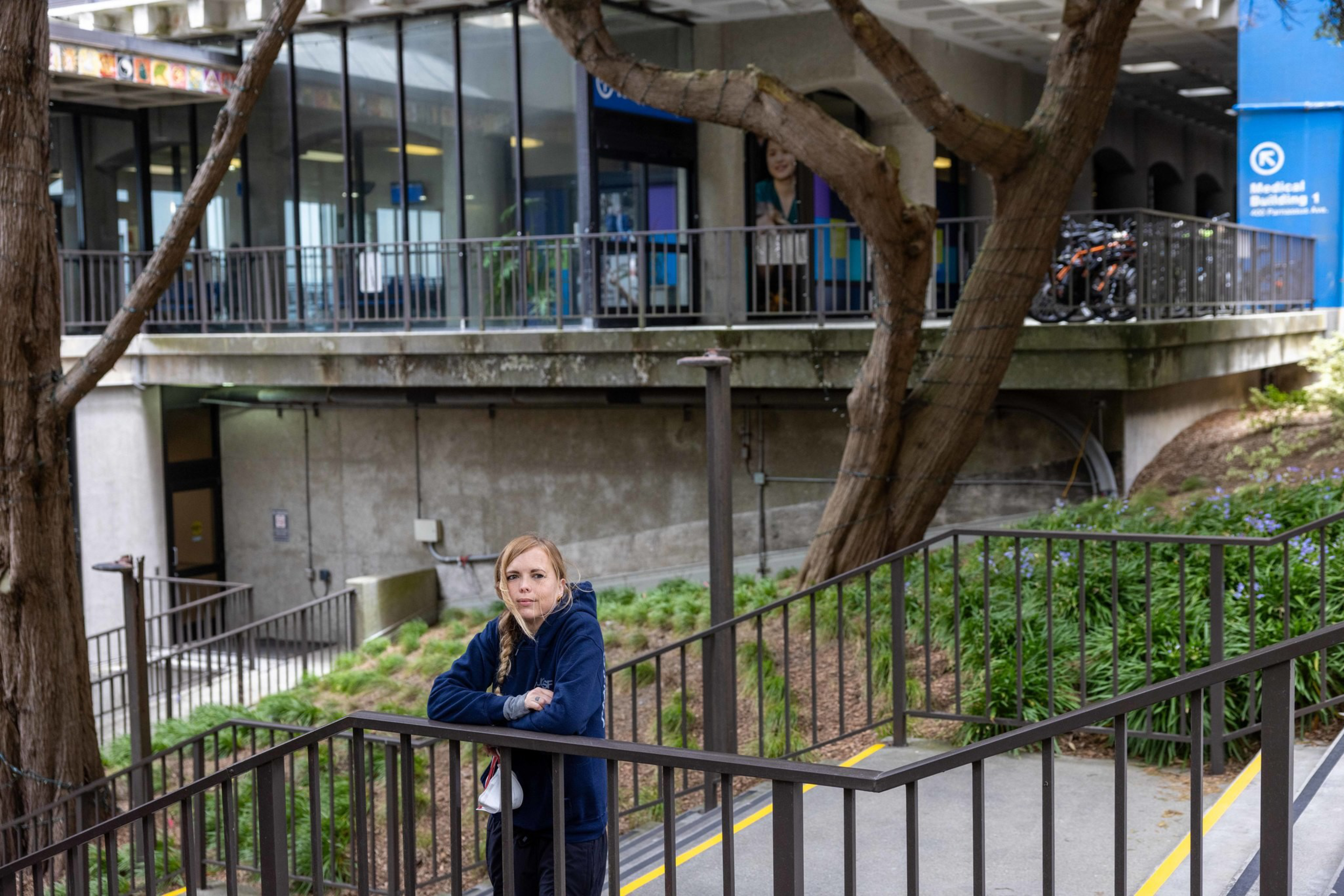 A woman in a blue hoodie leans on a black metal railing with trees, greenery, and a concrete building in the background.