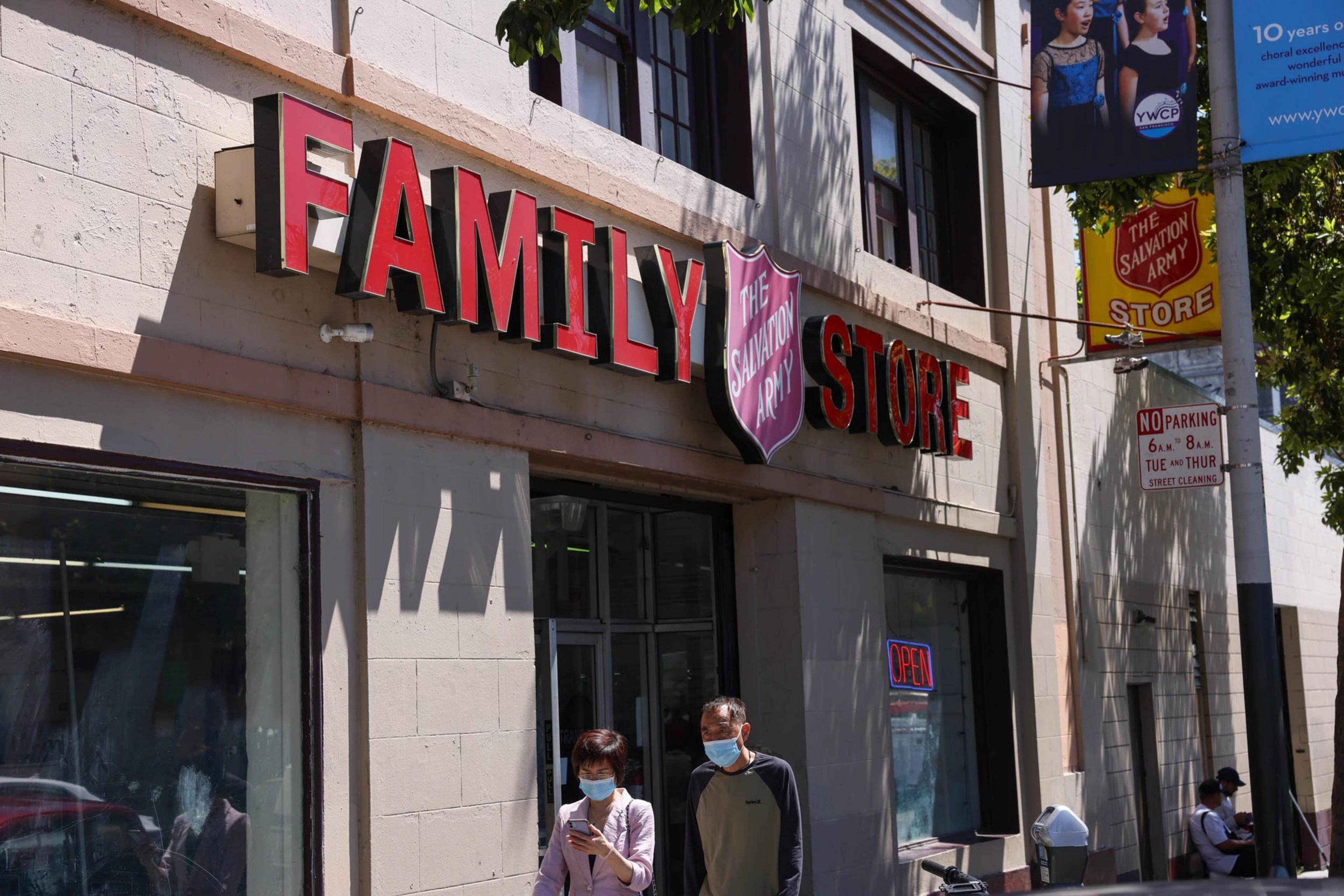 A Salvation Army Family Store storefront has red letters, a shield logo, and an "Open" sign, with two masked people walking outside.