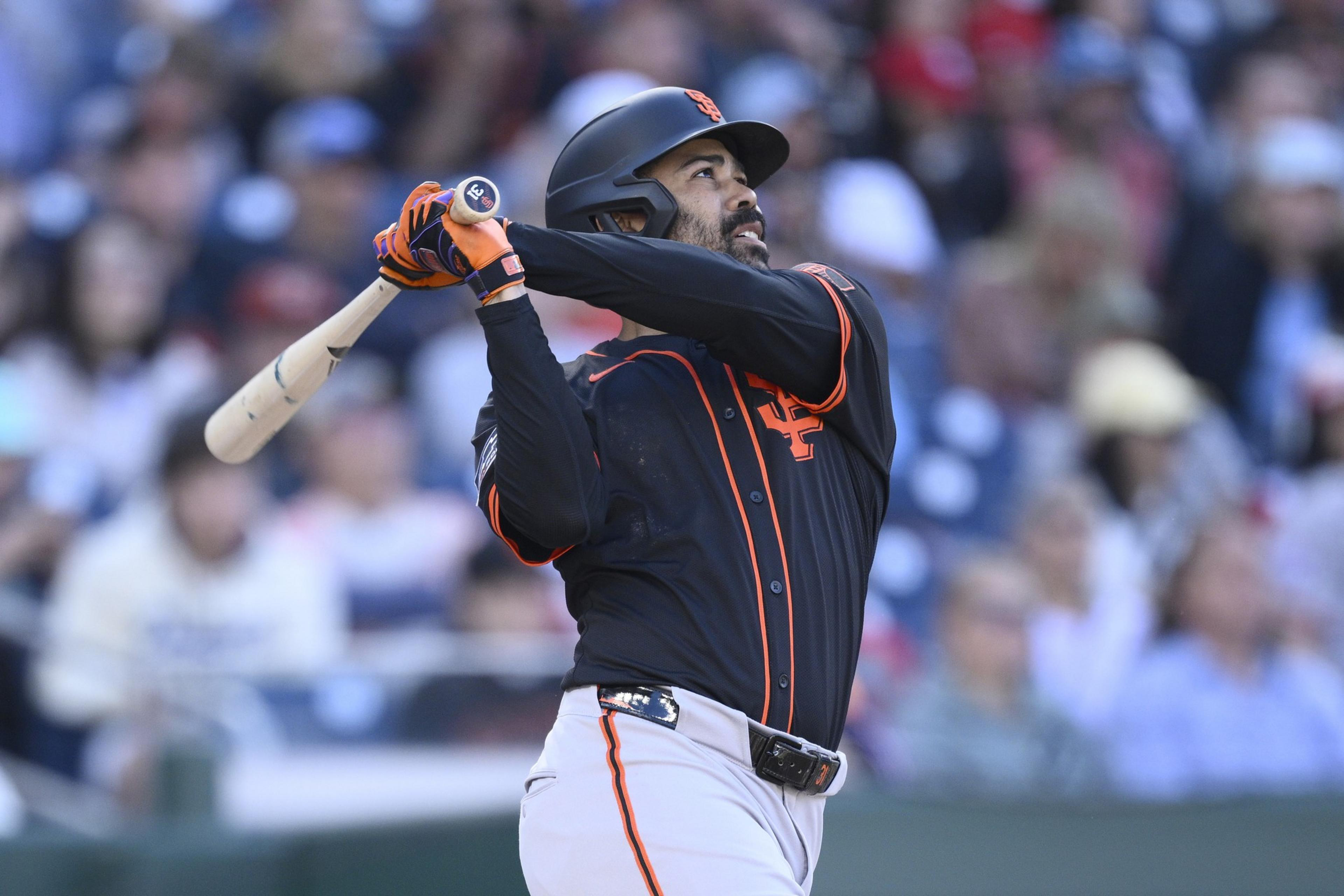 A baseball player in a black and orange uniform swings a bat during a game. He wears a helmet and gloves. The background shows a blurred crowd.