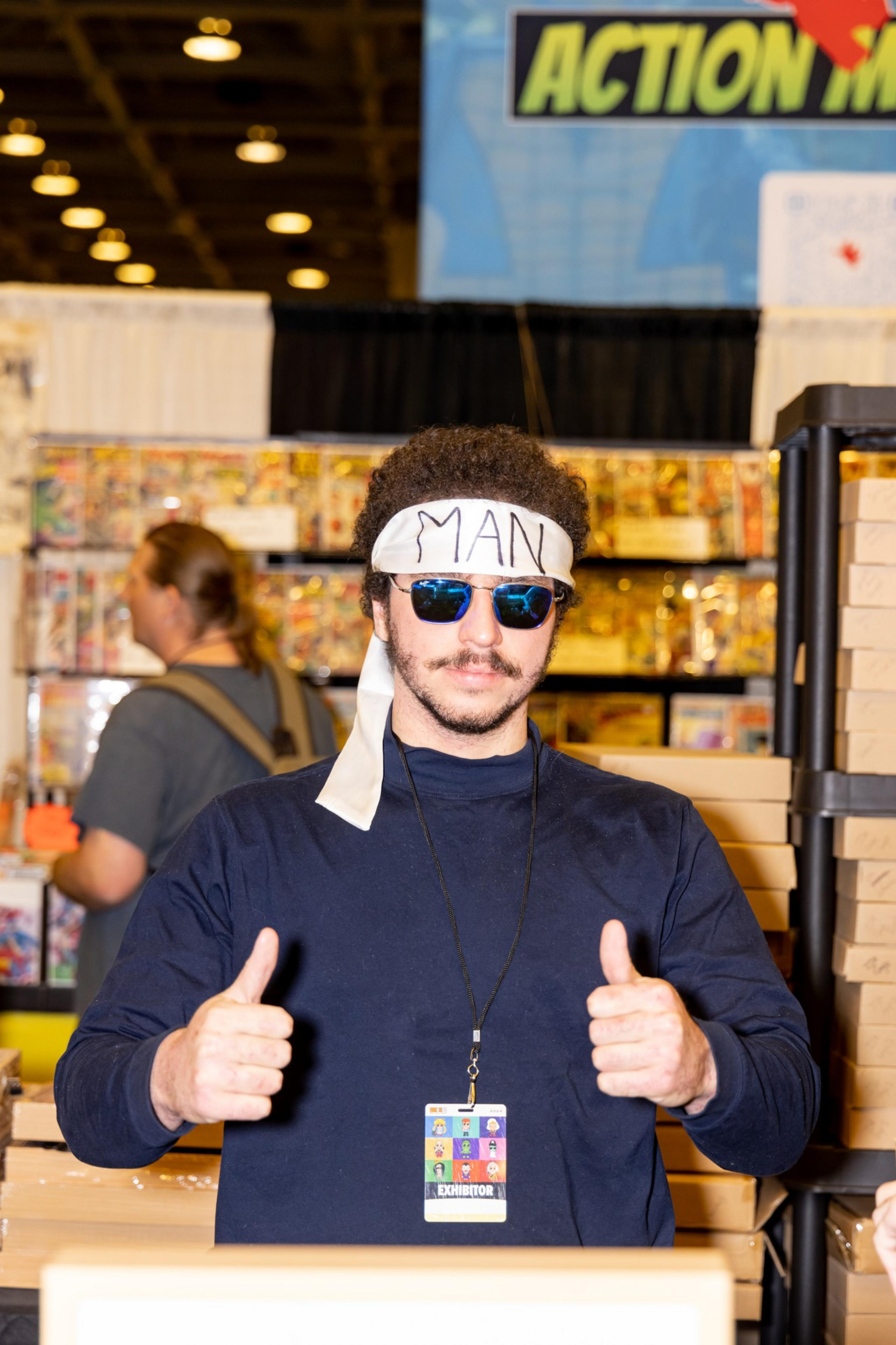 A man with a headband labeled &quot;MAN&quot; and sunglasses gives two thumbs up in a convention setting, wearing an exhibitor badge and standing near display shelves.