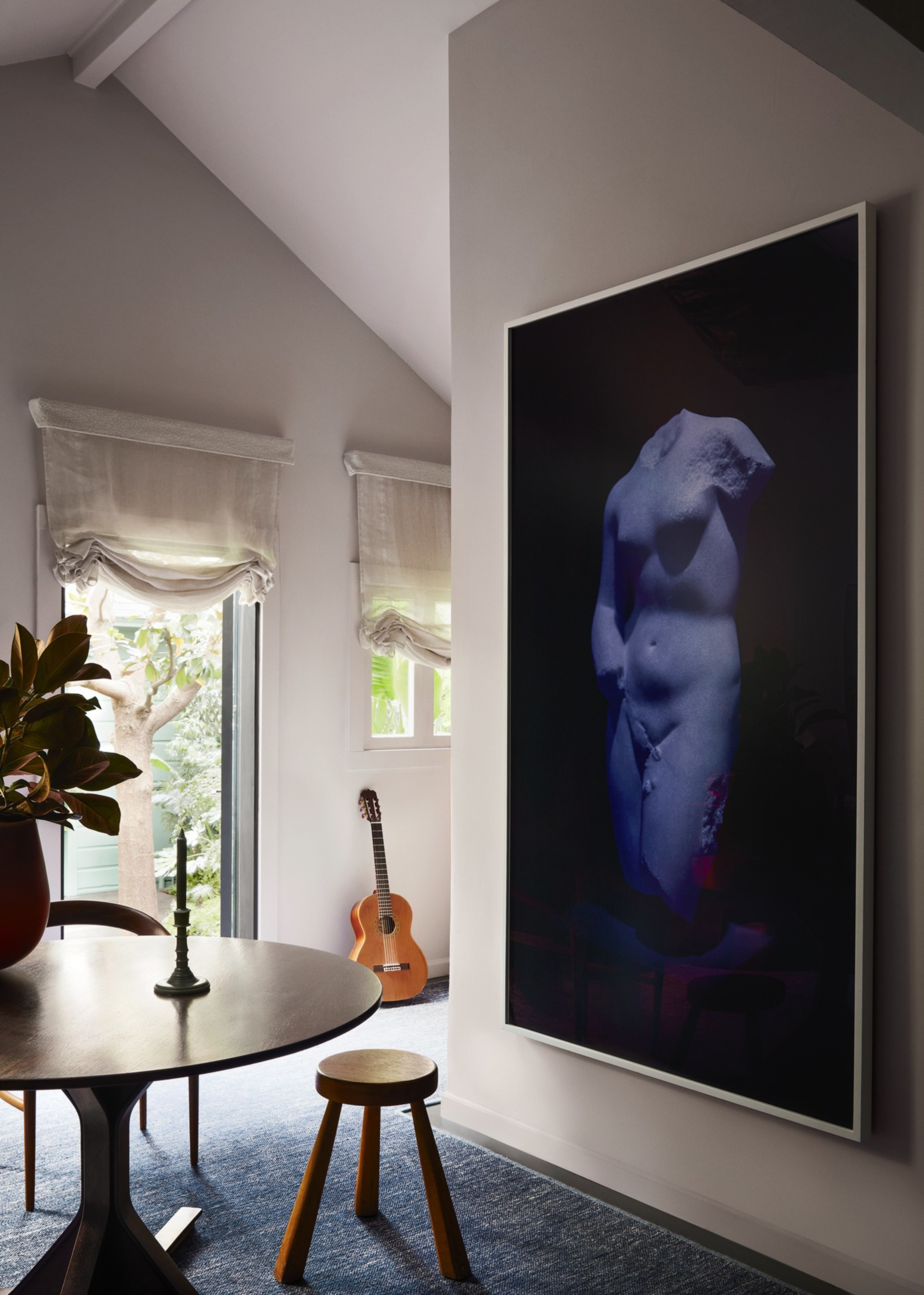 A room corner with a round wooden table, a wooden stool, a guitar by the windows, and a large framed photograph of a headless statue on the wall.