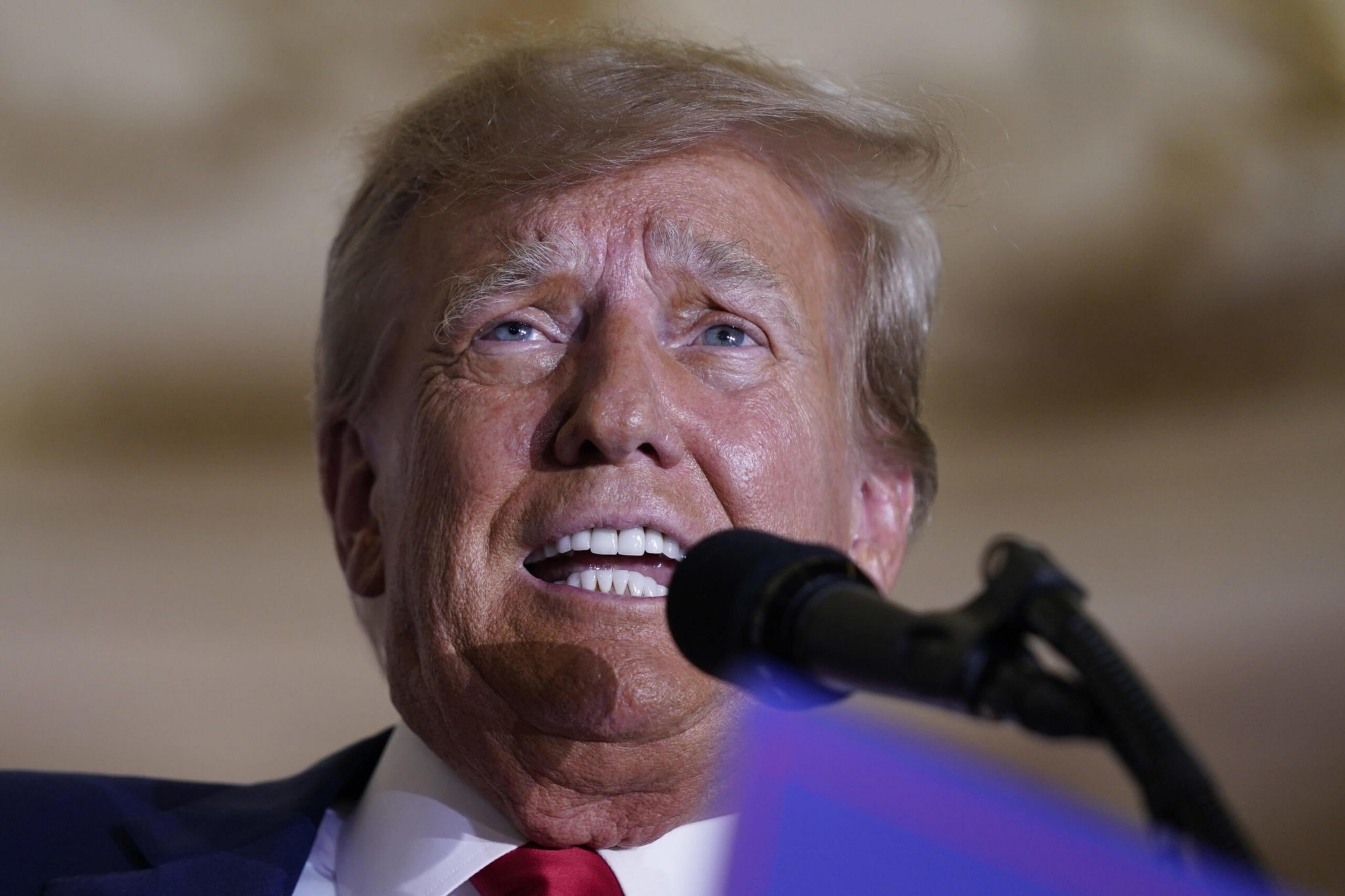 A person is speaking into a microphone, wearing a suit and red tie. The lighting highlights their facial details, and they appear to be looking upward.