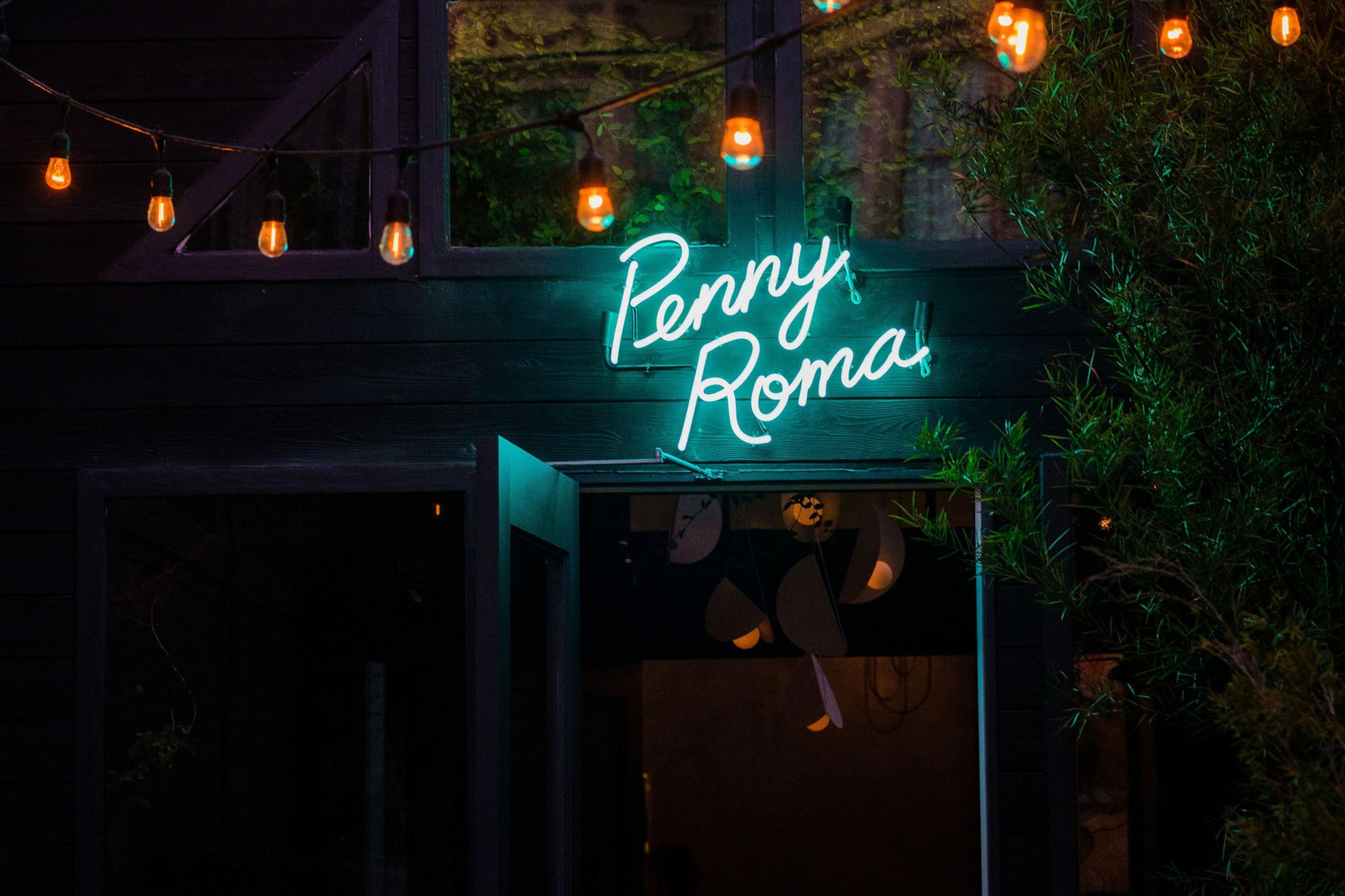 A teal neon sign reading "Penny Roma" hangs above an open doorway with string lights glowing warmly above it.