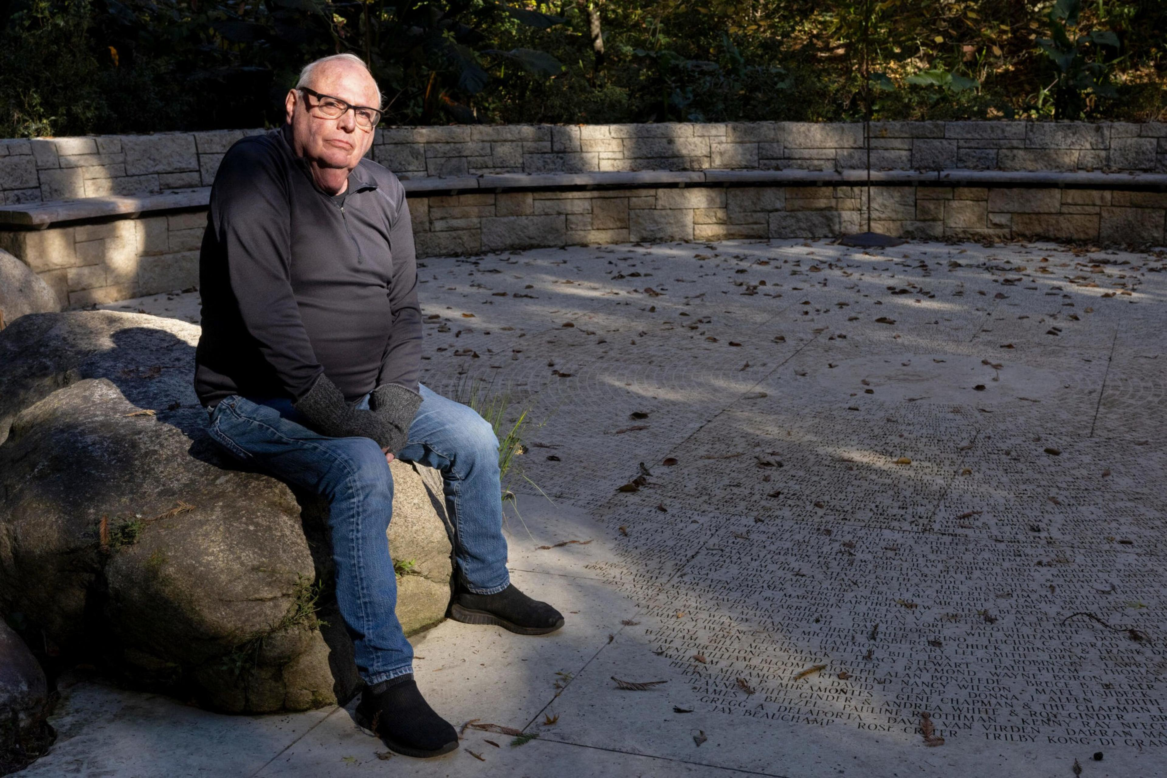 An older man wearing glasses, a black long-sleeve shirt, blue jeans, and gloves sits on a large rock beside a stone bench in a shaded outdoor area.