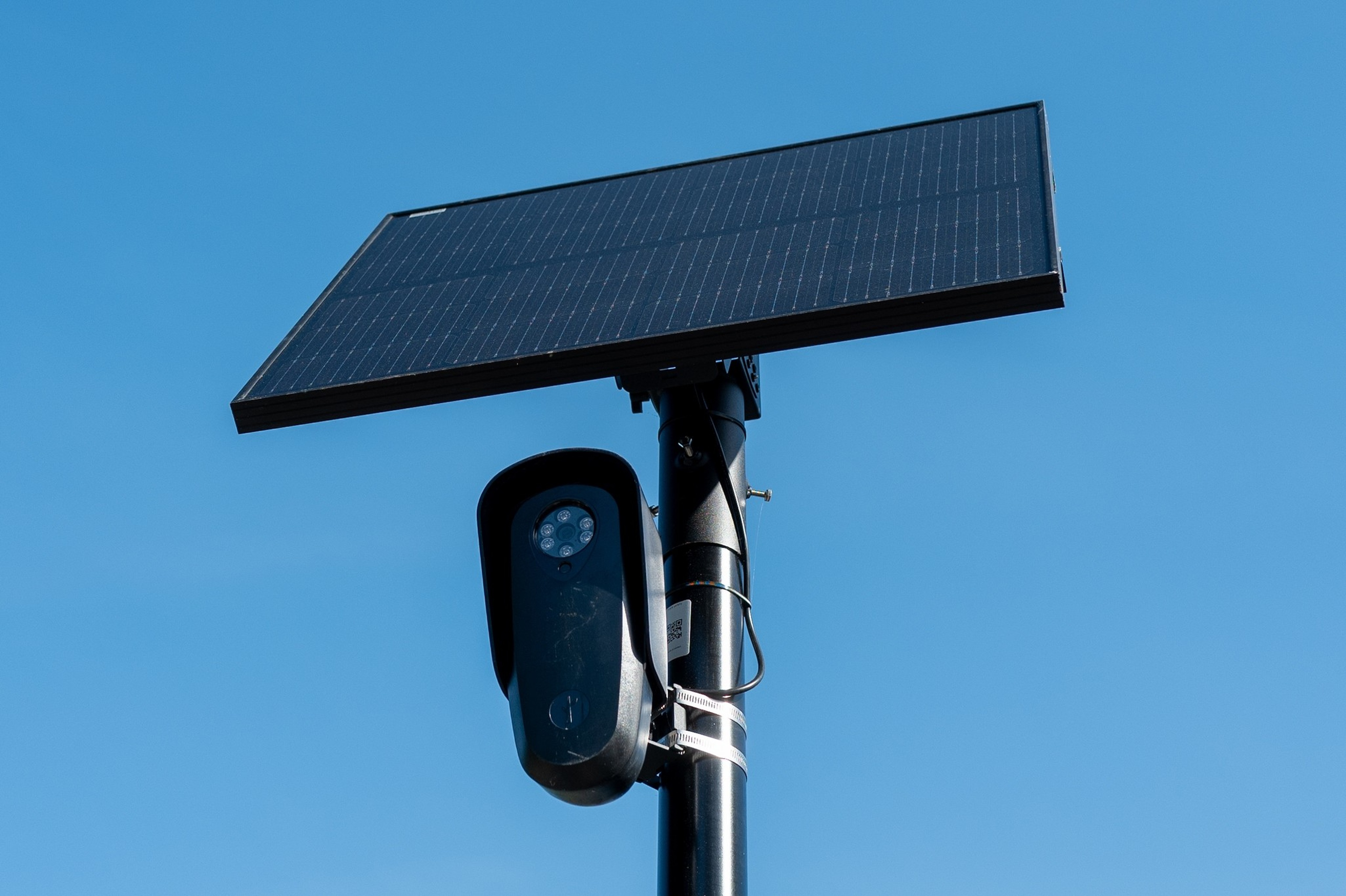 A black security camera is mounted on a pole beneath a large solar panel against a clear blue sky.