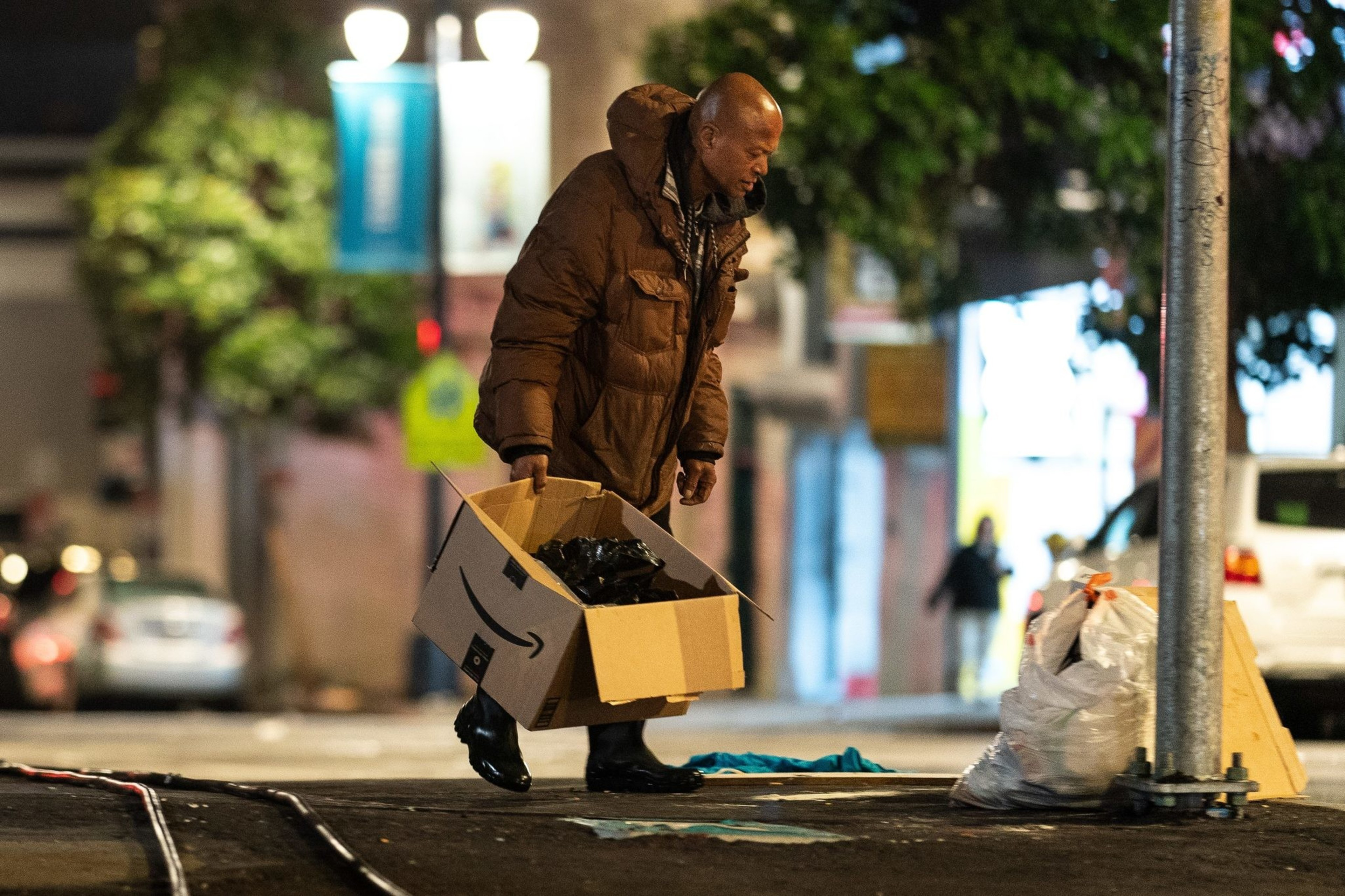 A person with a brown coat walks in the street with a box in hand.