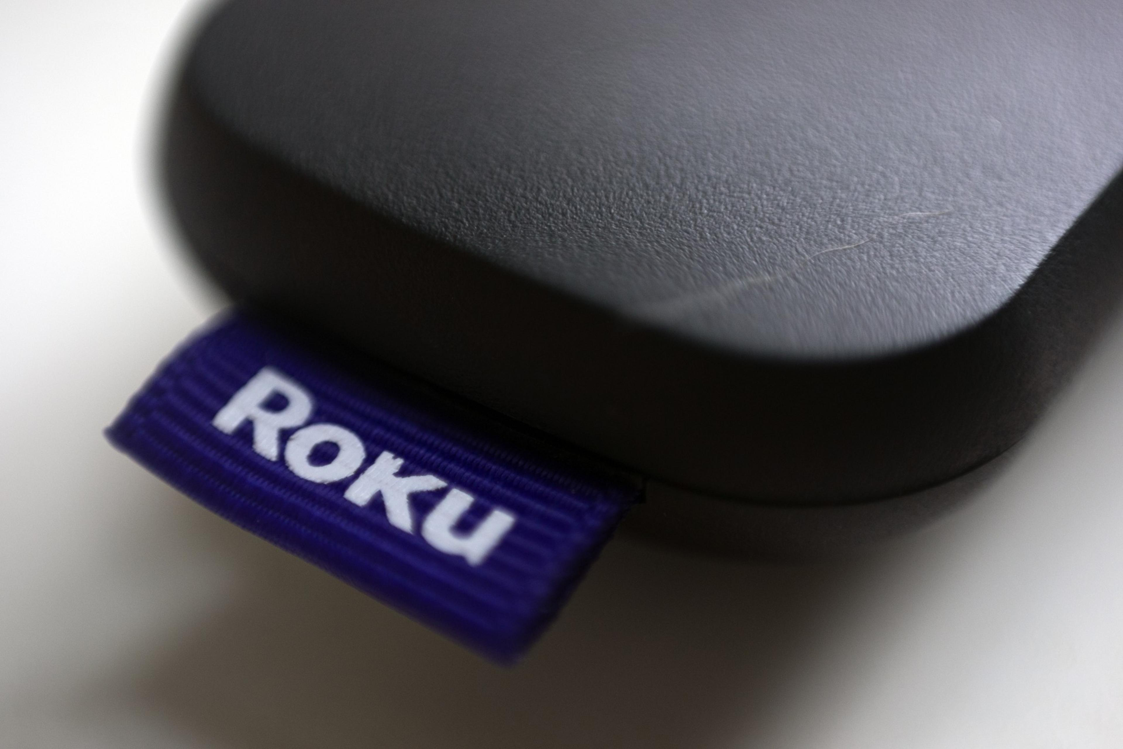A close-up of a black Roku streaming device with a purple fabric tag displaying the white Roku logo attached to its side.