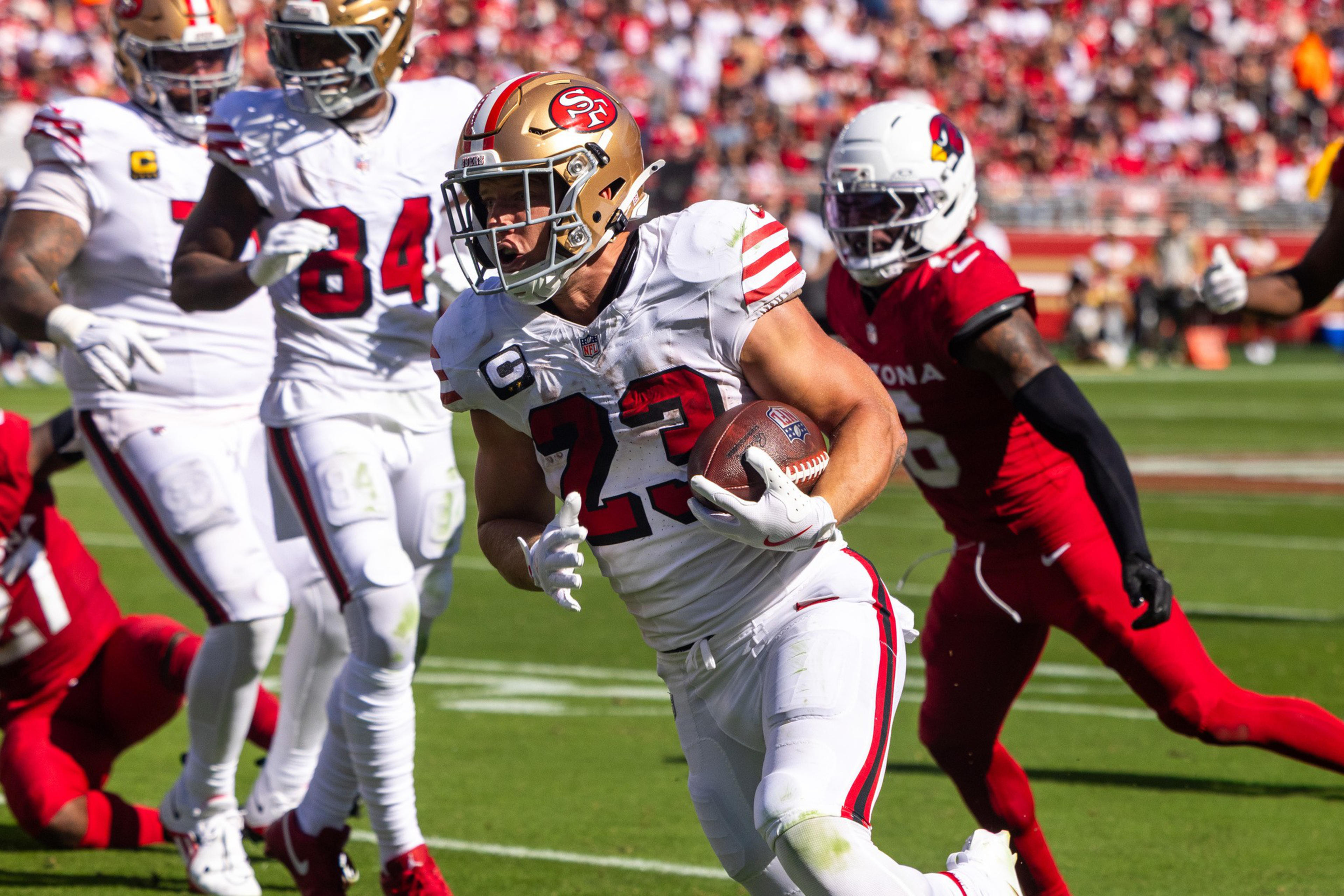 A San Francisco 49ers player in white runs with the football while players from the Arizona Cardinals in red try to catch him.