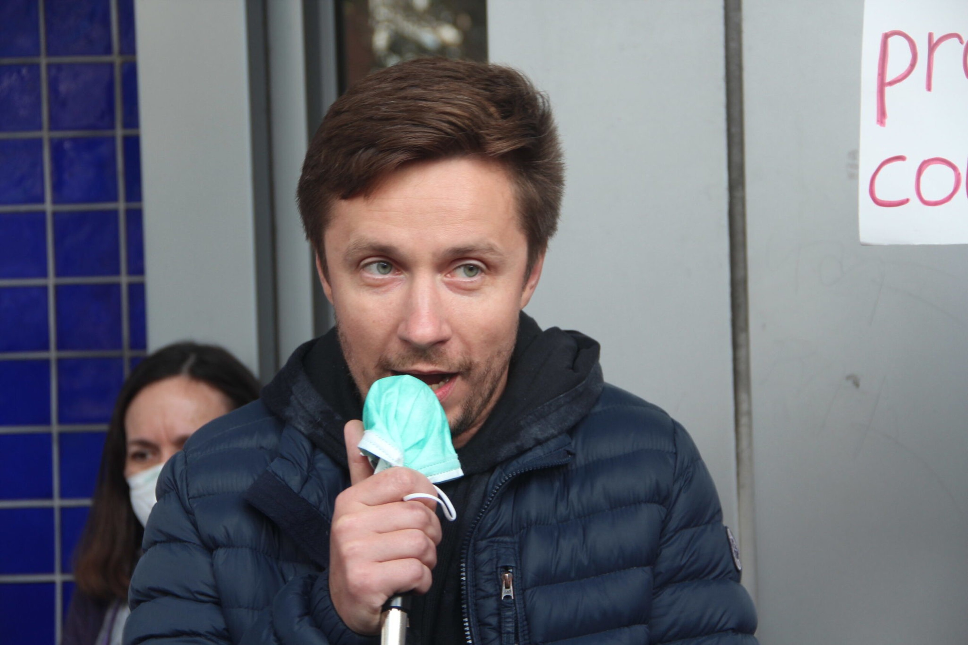 A man in a blue jacket holds a microphone covered with a green mask near his mouth, with a woman wearing a mask visible behind him.