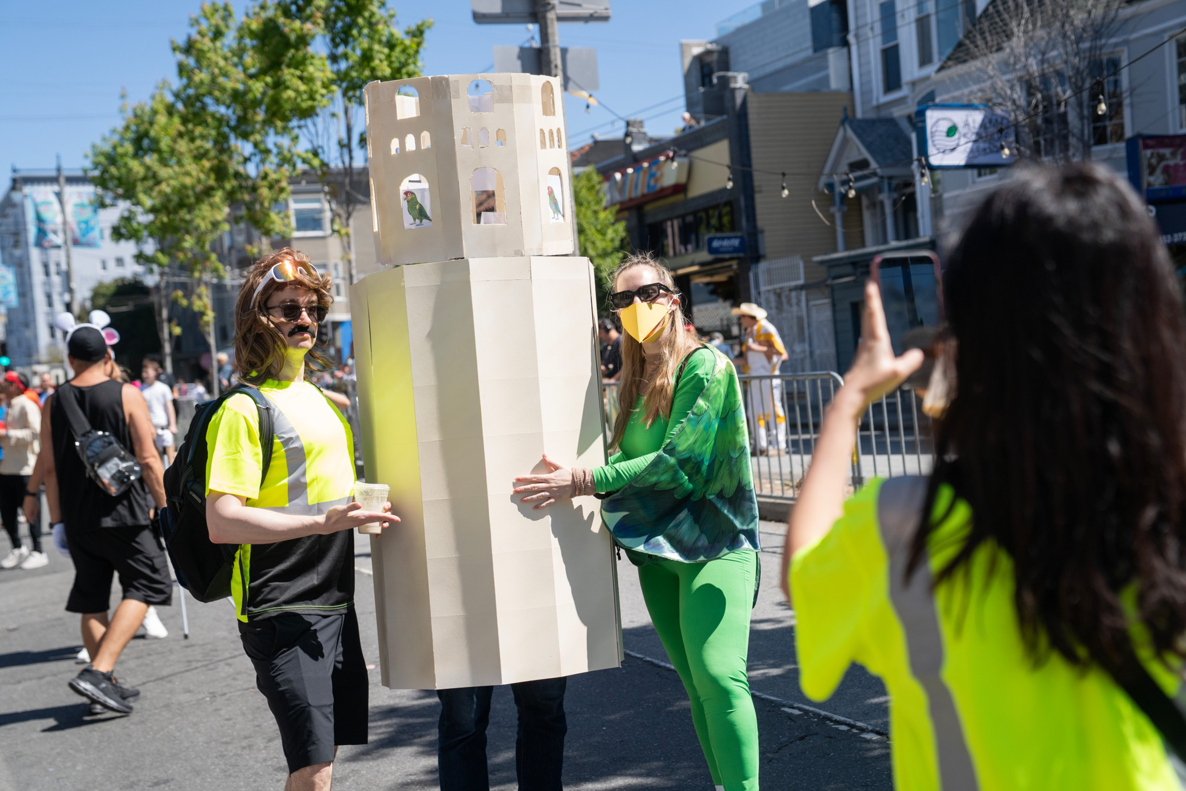 Two people pose, one inside a tall beige paper tower costume with bird cutouts, while another takes their photo on a sunny street with pedestrians.