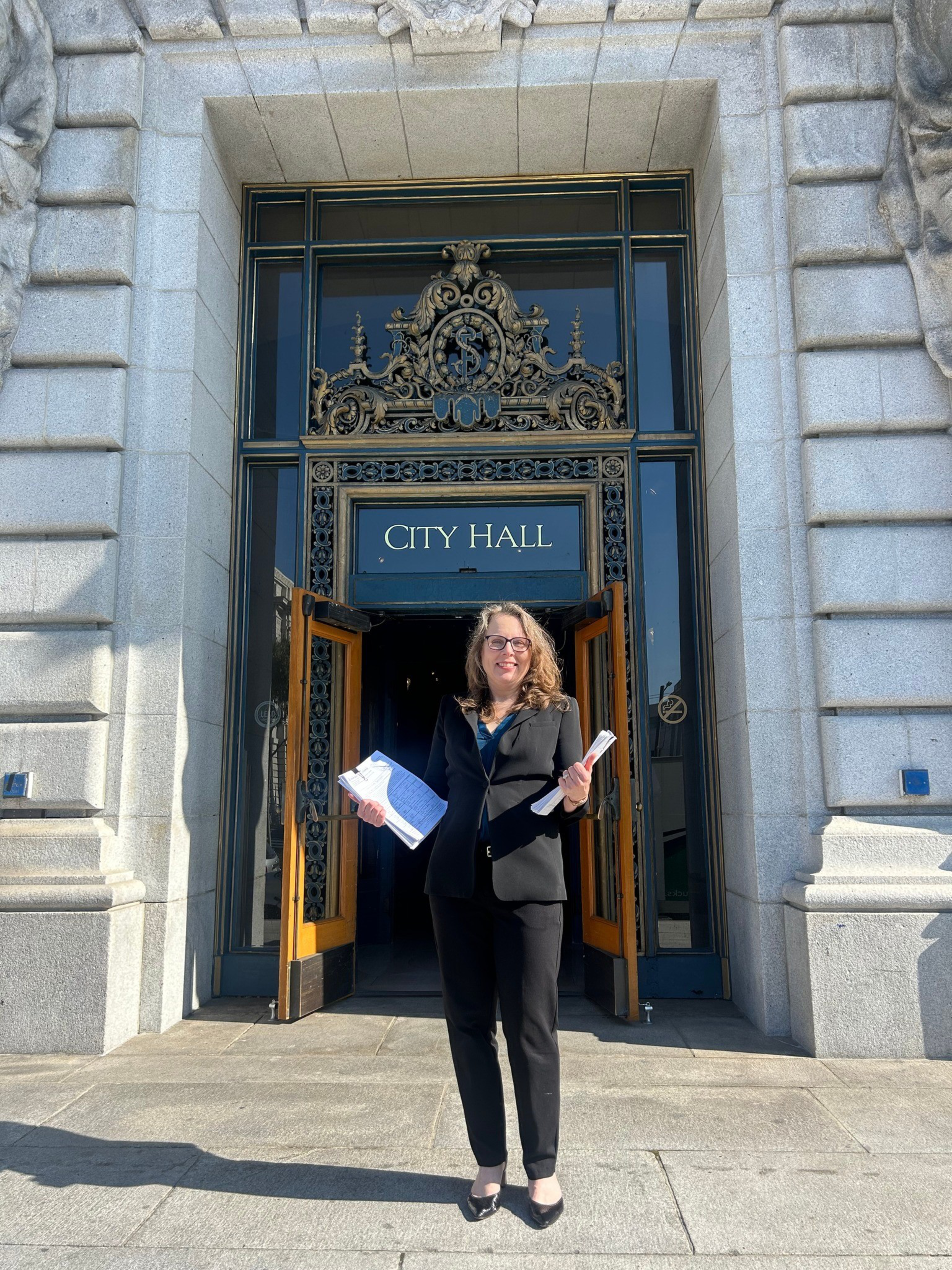 A woman in a black suit stands smiling with papers in both hands outside the open doors of a stone building labeled “City Hall.”