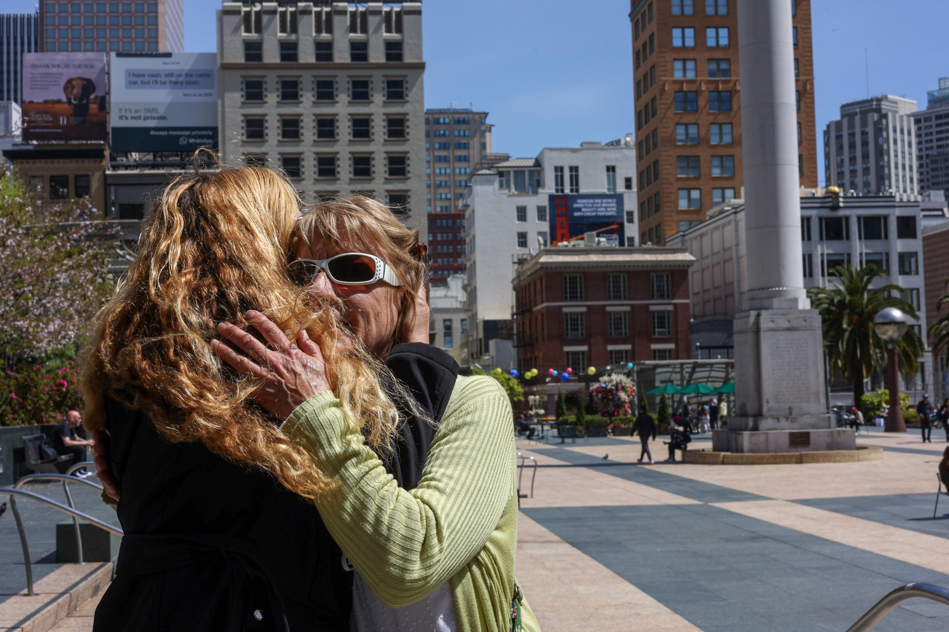 Two women embrace closely in an urban plaza surrounded by tall buildings and a column monument under a clear blue sky.