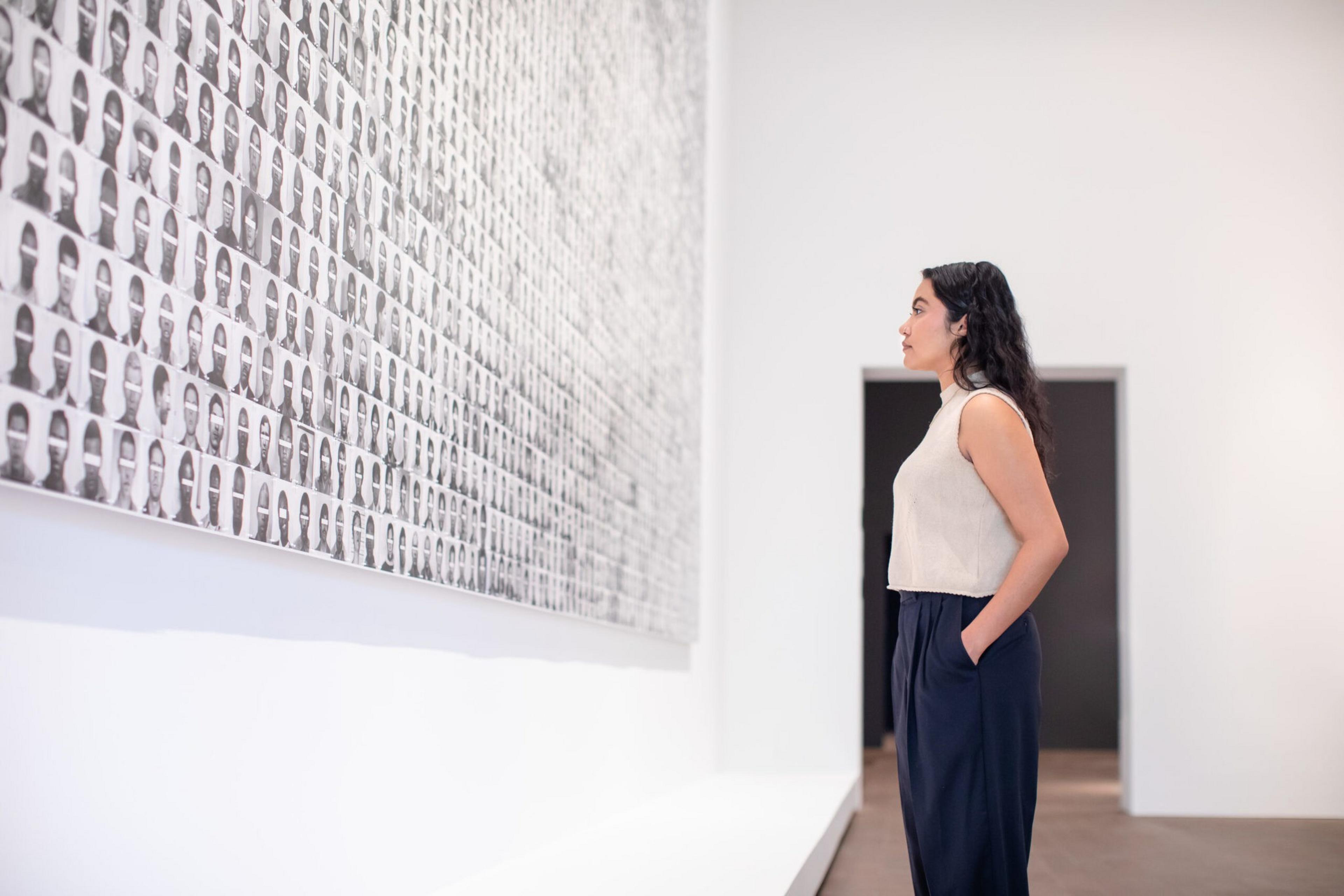 A woman stands in a gallery, closely observing a large wall covered with numerous black-and-white portrait photos arranged in a grid.