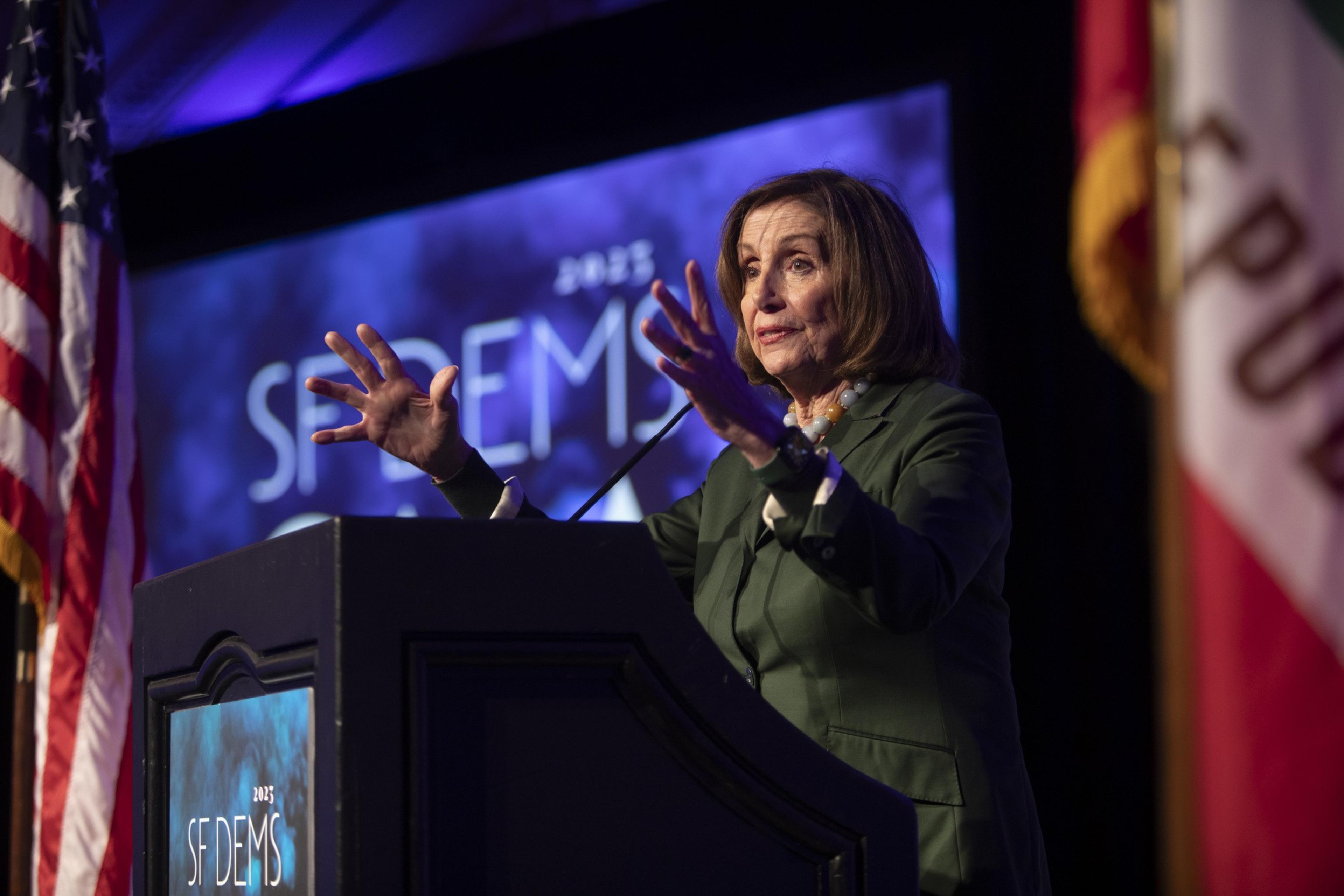 A woman in a green suit speaks passionately at a podium with &quot;2023 SF DEMS&quot; on it, flanked by the American and California flags. A purple-lit backdrop is behind her.