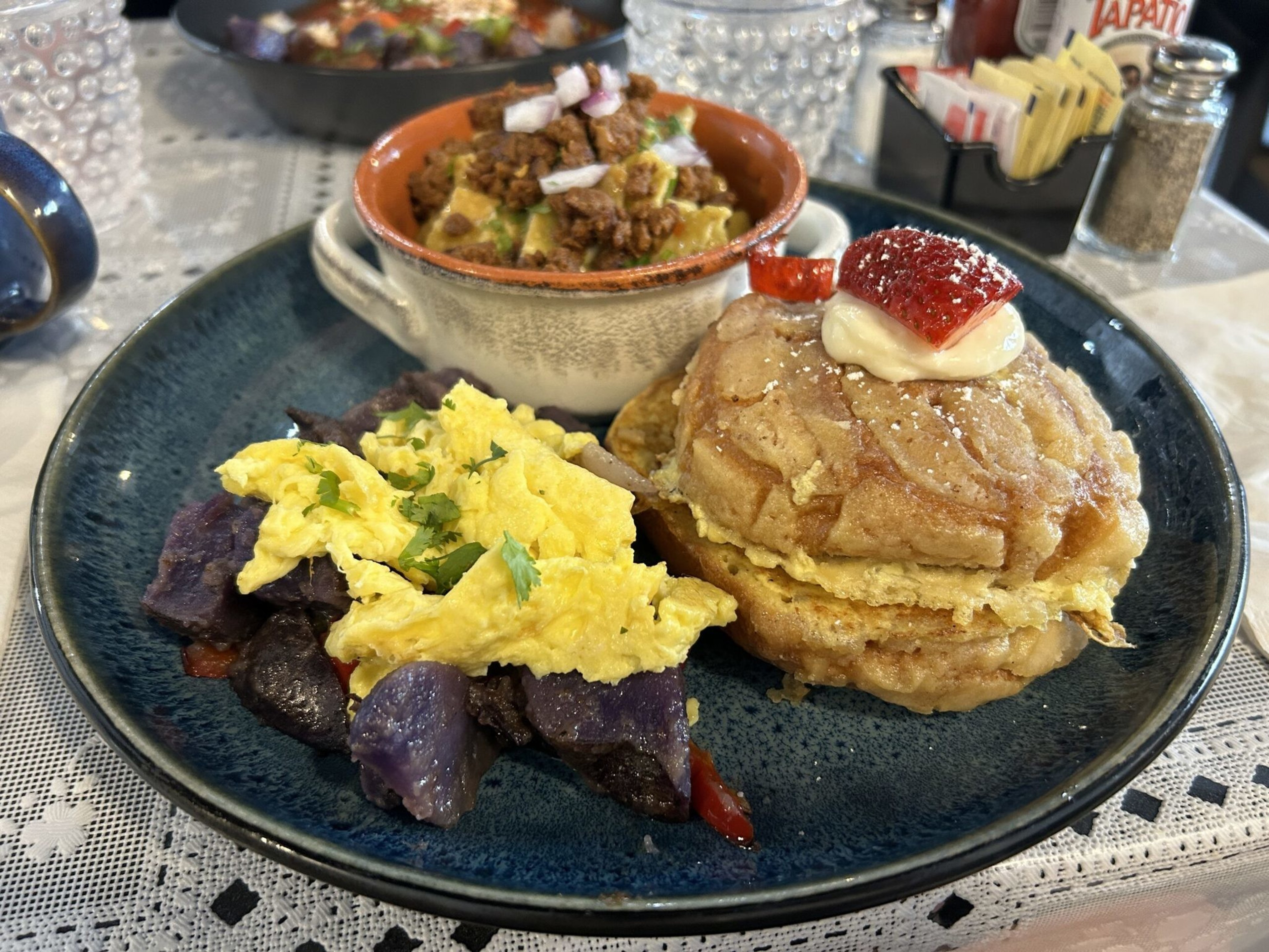 Scrambled eggs over purple potatoes, two stacked pancakes topped with cream and a strawberry slice, and a cup of seasoned ground meat with chopped onions.