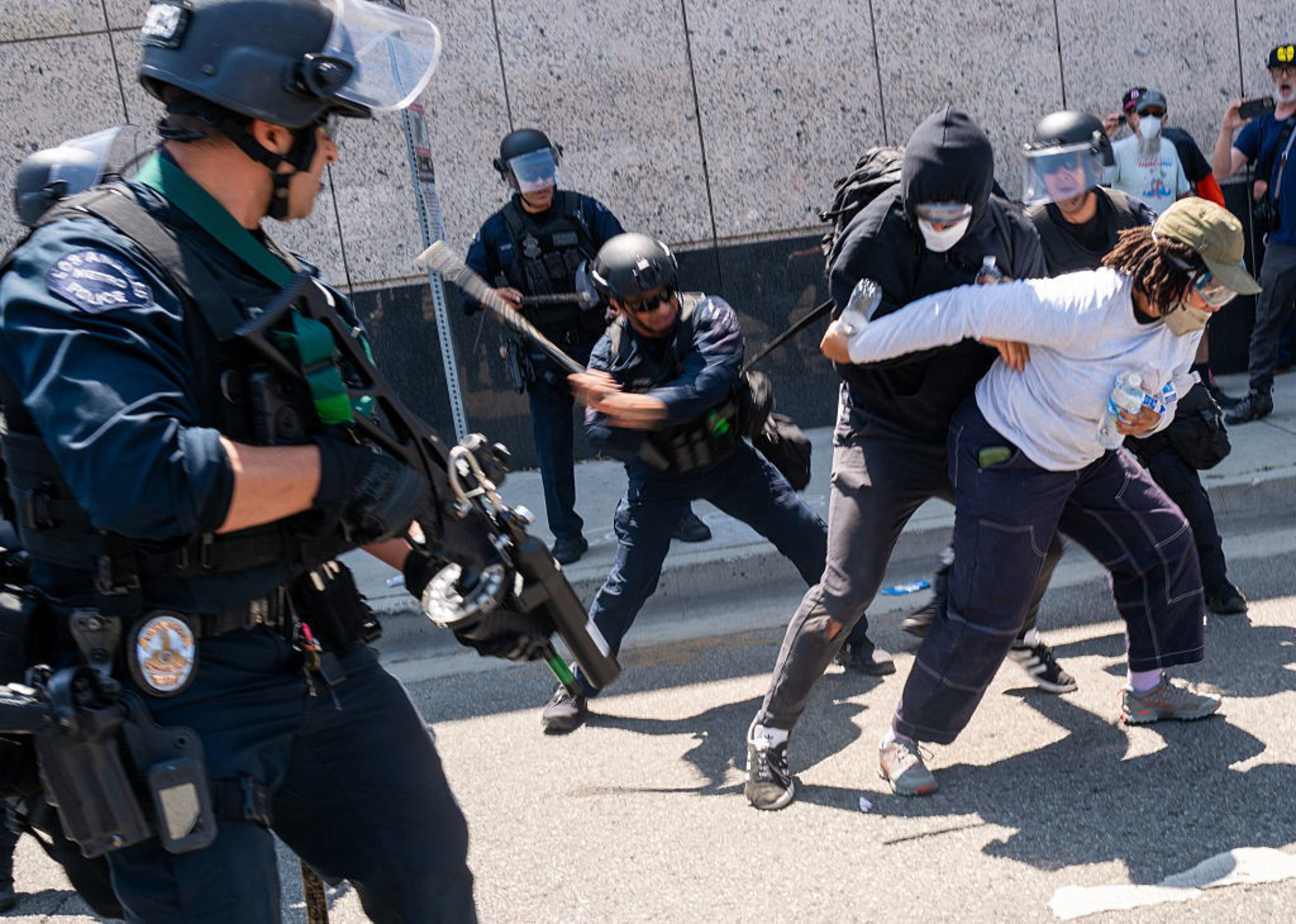Police officers in riot gear confront protesters on a street. One officer wields a baton, while protesters appear to struggle. Tension is evident.