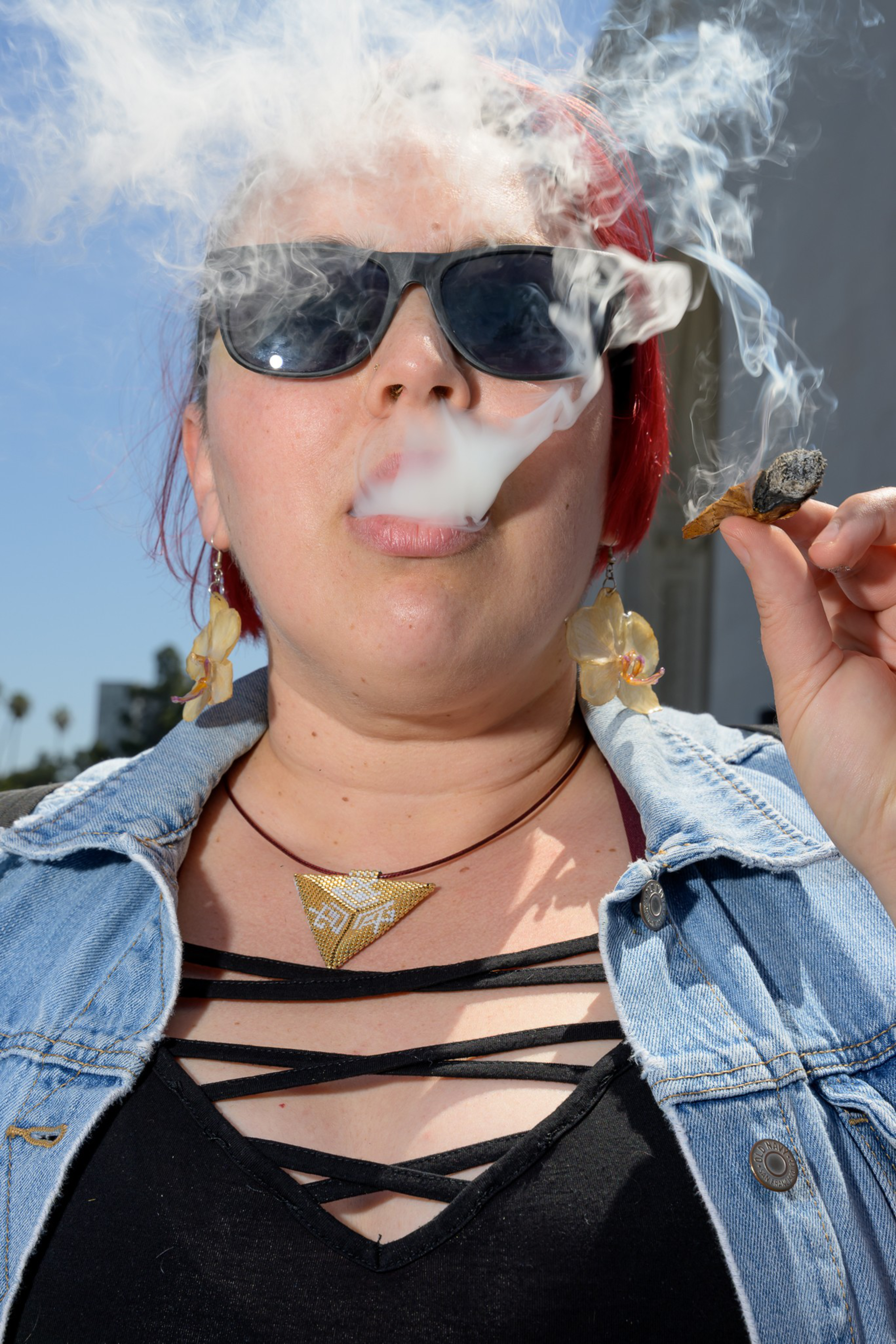 A person with red hair, wearing sunglasses and a denim jacket, is exhaling smoke from a small pipe. They have a beaded necklace and large flower earrings.