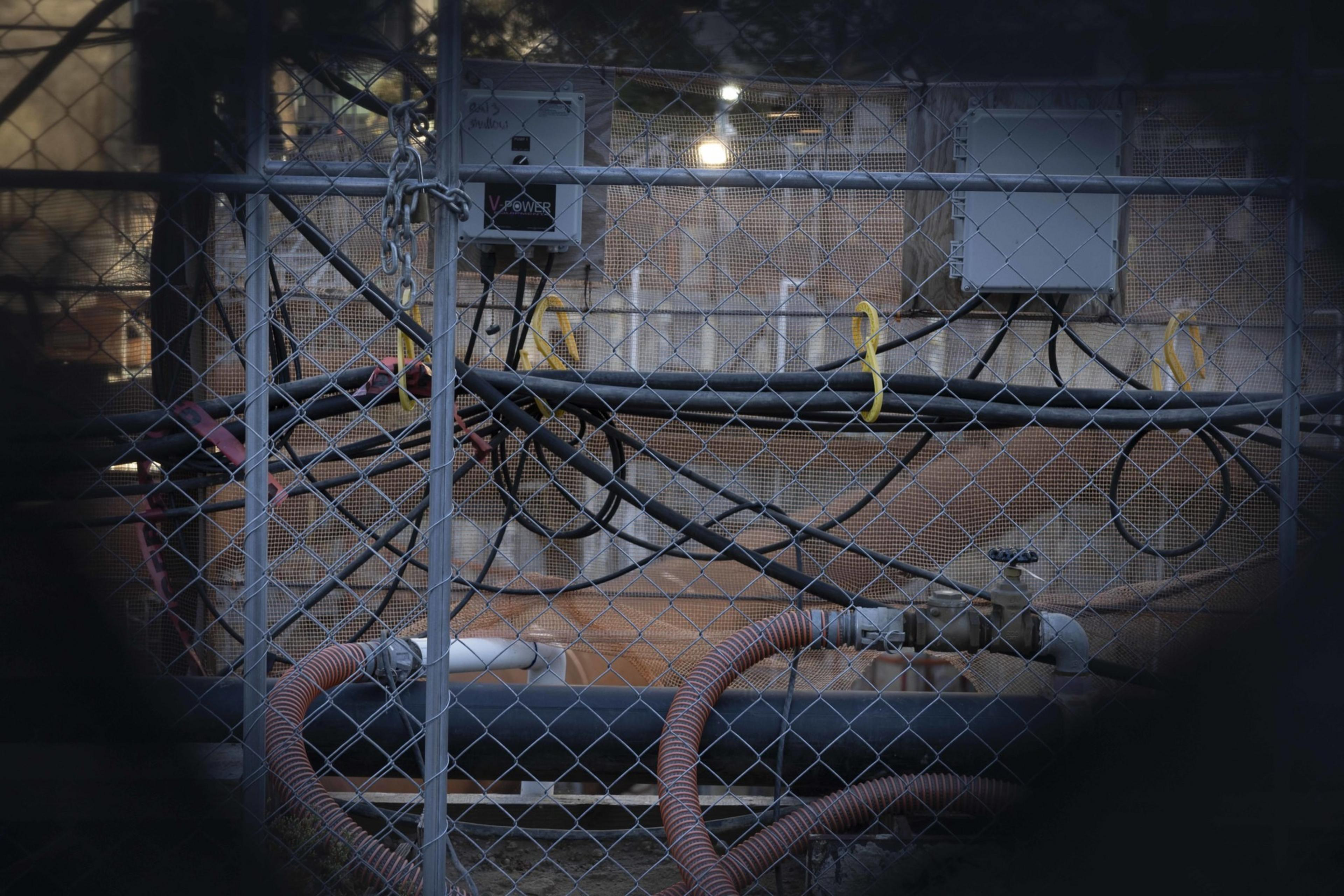 The image shows a construction area enclosed by a chain-link fence. Inside are electrical boxes, cables, and pipes, with various wires and tubes connected.