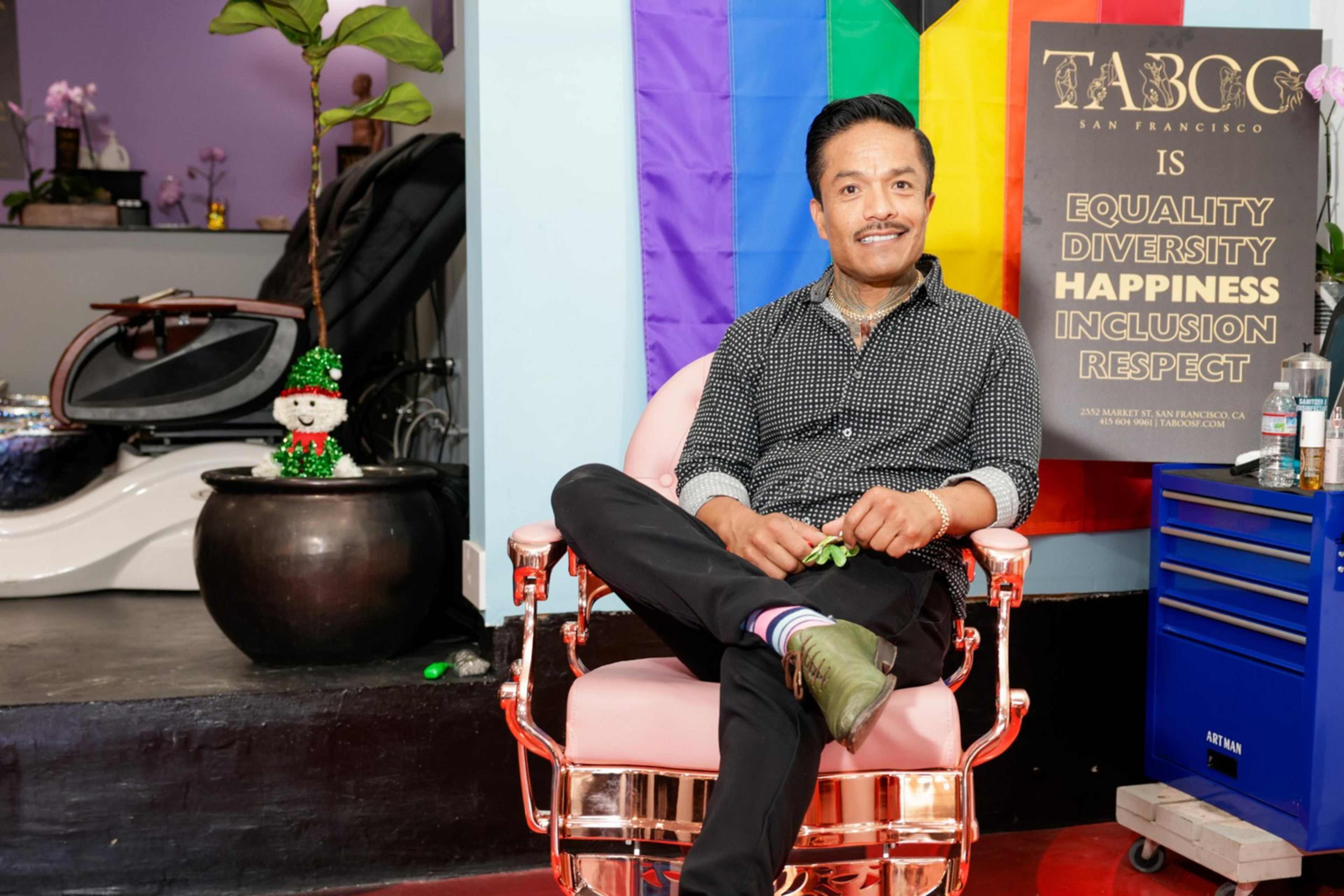 A person sits cross-legged in a barber chair, smiling. Behind them is a sign promoting equality and diversity, and a colorful flag. The environment is bright and welcoming.